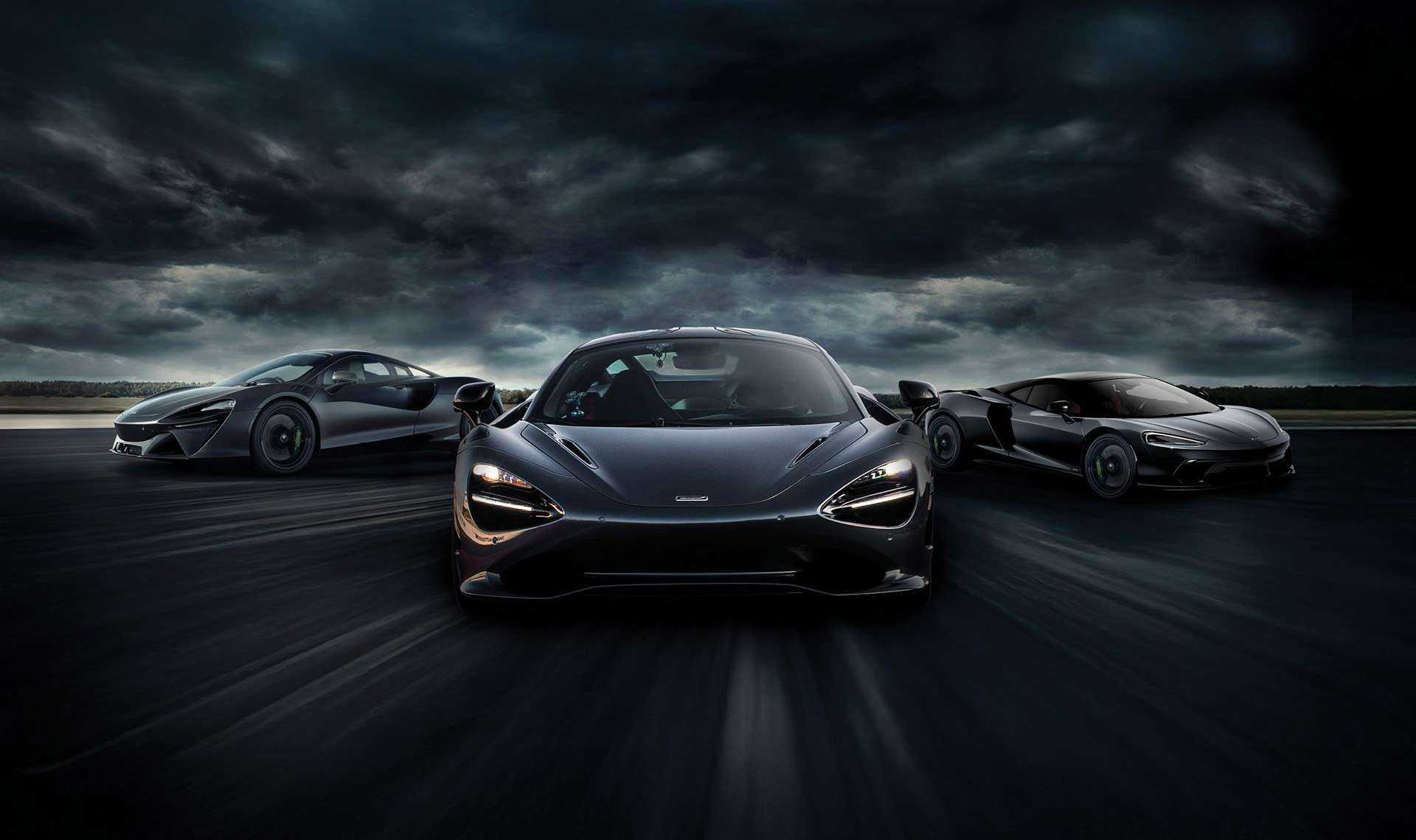 A dynamic, wide-angle shot of three dark McLaren supercars on a blurred track, with the central car prominently in the foreground, under a dramatic, cloudy sky.
