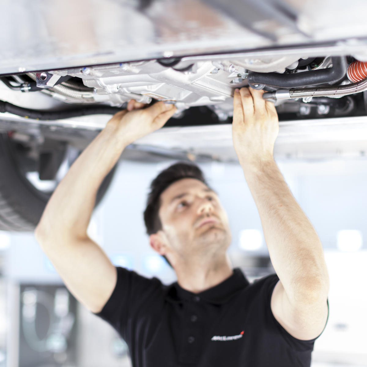 A McLaren technician in a black polo shirt with the brand's logo works on the undercarriage of a car, focusing on a specific component.
