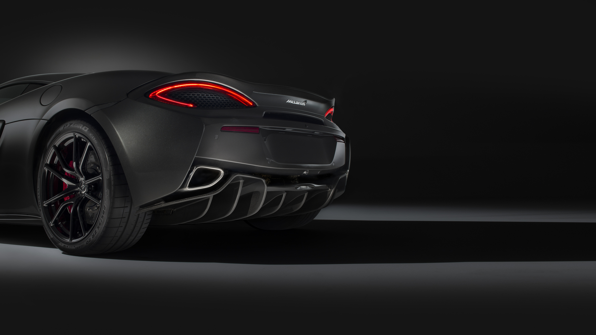 A close-up, rear-three-quarter view of a dark grey McLaren sports car in a studio setting with a black background. The image focuses on the curved taillight, aerodynamic diffuser, and exhaust pipes.