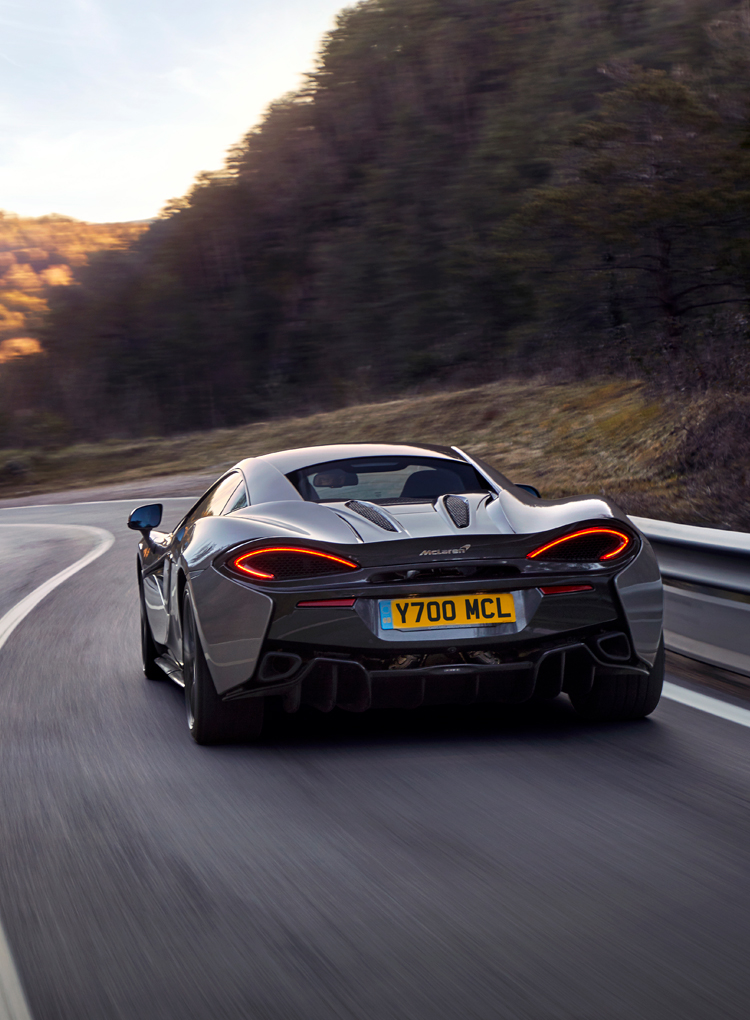 A grey McLaren 570S driving around a bend at speed, surrounded by trees.