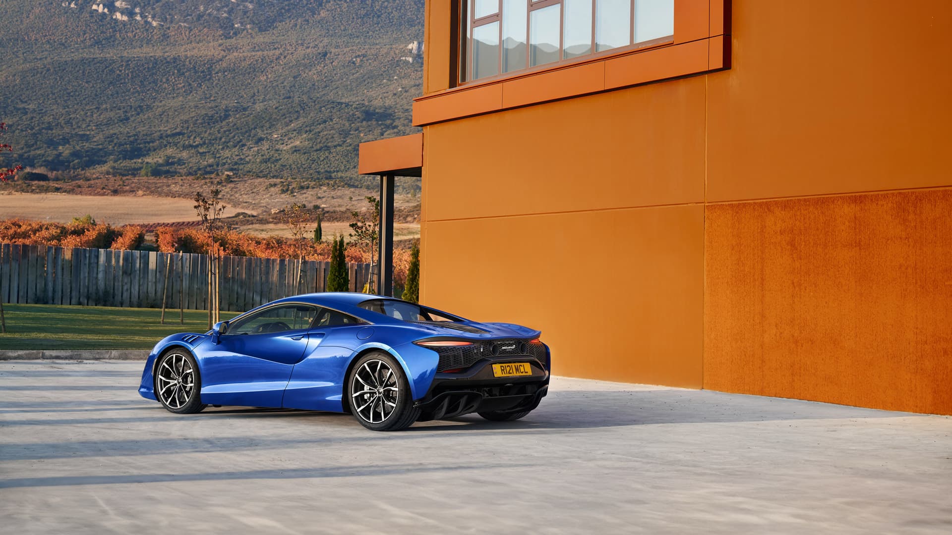 Rear shot of blue McLaren Artura parked in front of an orange building, in the mountains.