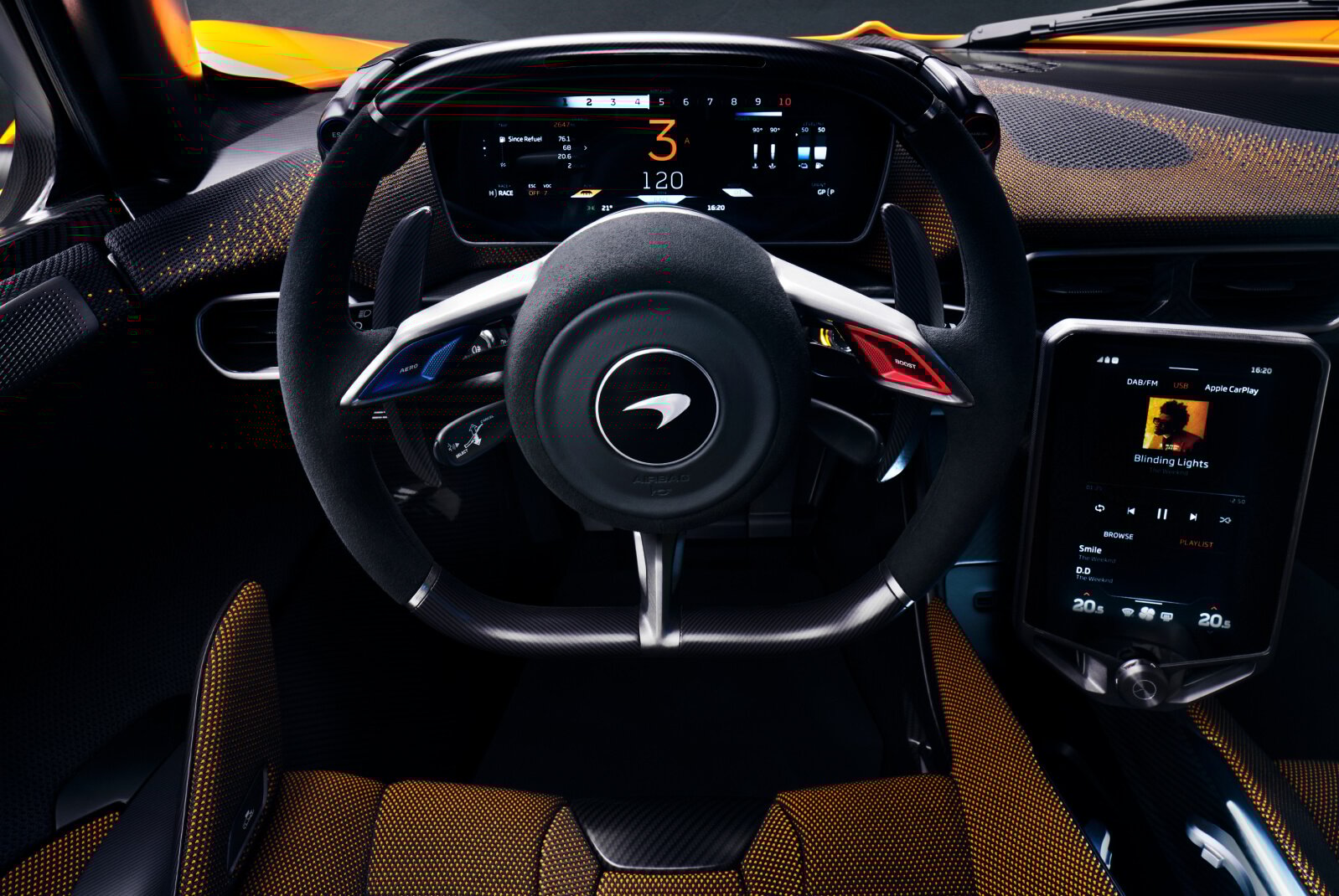 A dashboard of a McLaren W1 with a steering wheel, showing the driver display and the carbon fibre design of the steering wheel with shift lights and blue and red buttons.