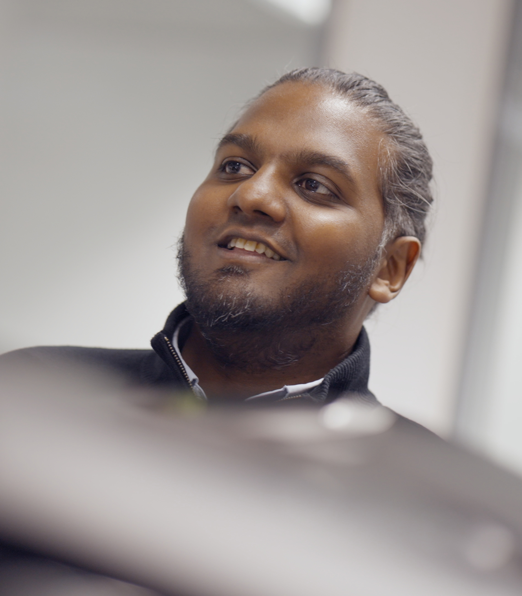 A professional headshot of a man with a beard and hair in a bun, smiling while looking up and to the side in a brightly lit office.
