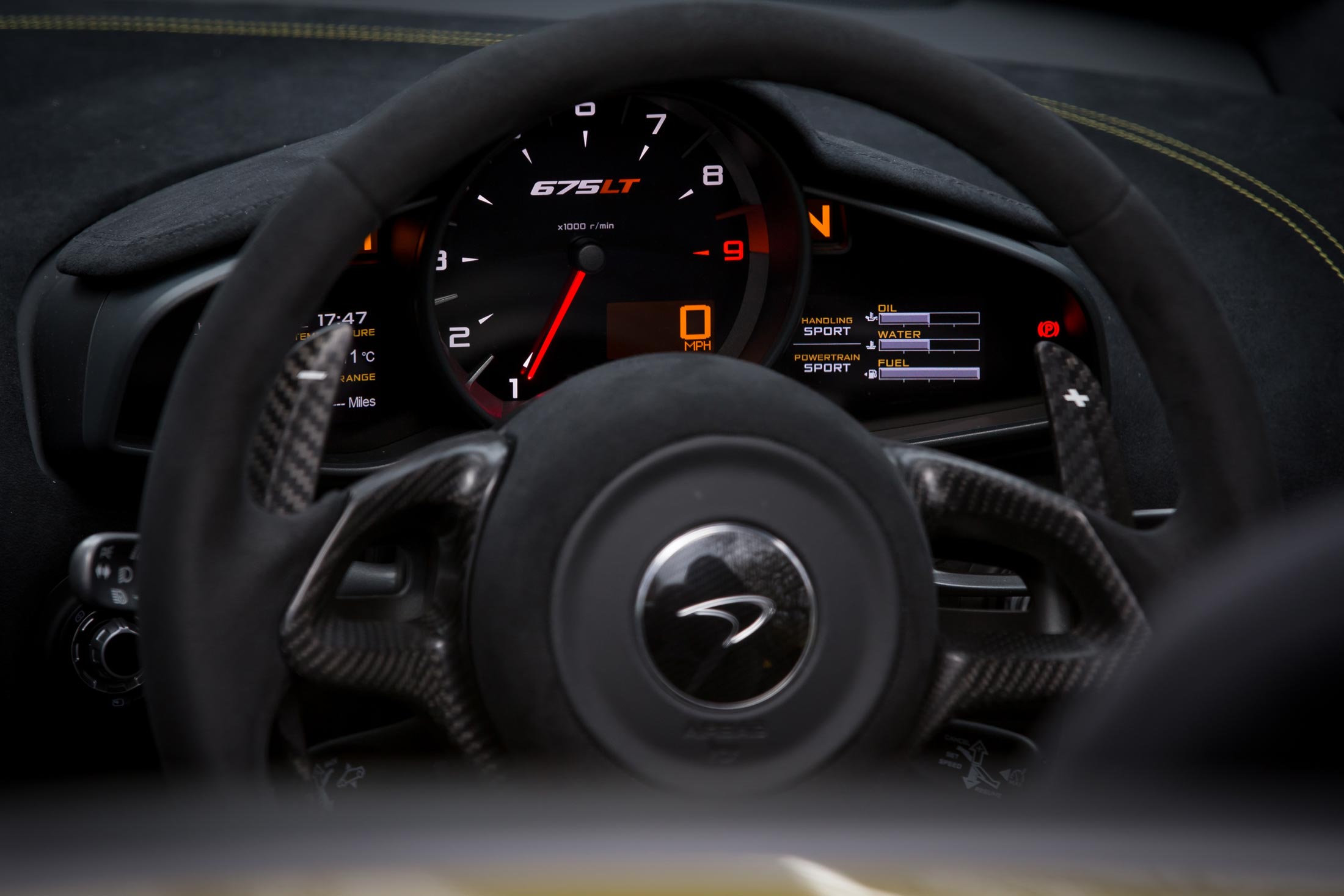 A close-up of the driver's perspective inside a McLaren 675LT, focusing on the digital dashboard with a red tachometer, the Alcantara steering wheel, and carbon fiber paddle shifters.