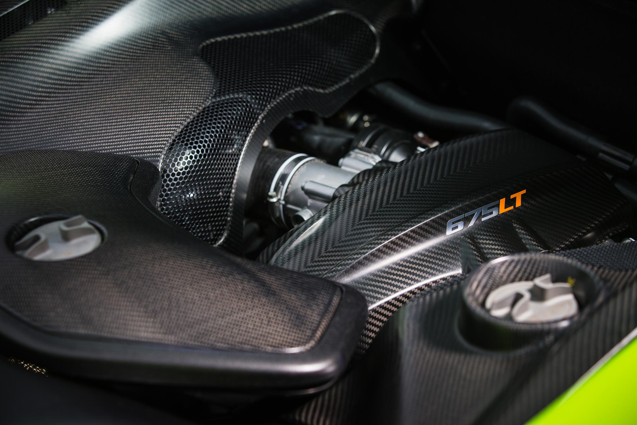 A close-up of a McLaren engine bay, highlighting the lightweight carbon fiber components with a '675LT' logo and the intricate mesh grille.