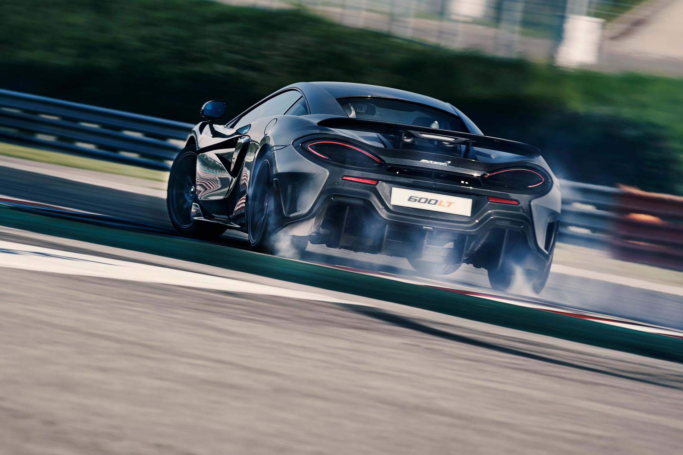A rear-three-quarter view of a dark grey McLaren 600LT drifting on a racetrack, with tire smoke trailing behind the rear wheels.