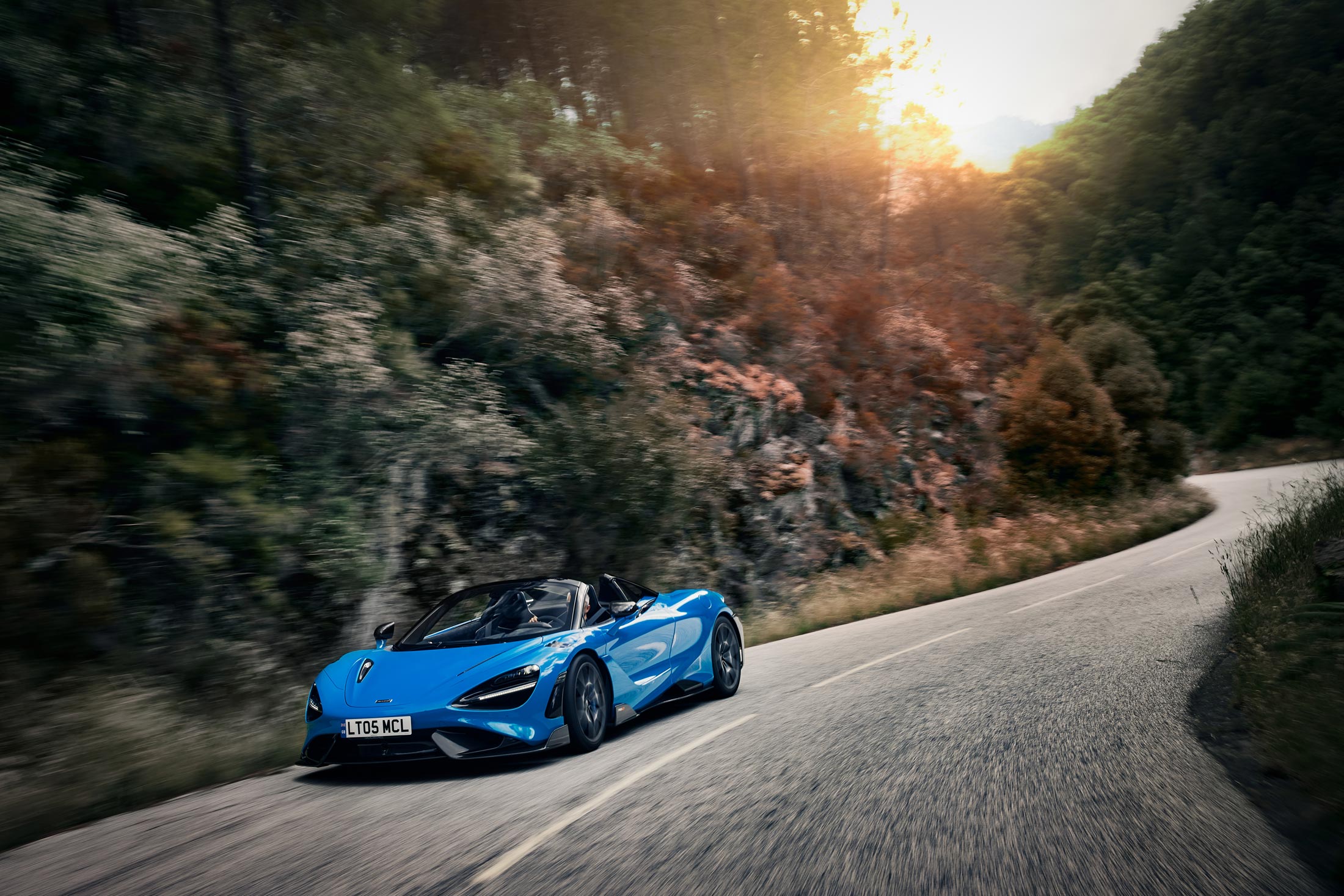 A vibrant blue McLaren 720S Spider is captured in motion on a sun-drenched, winding road with a forest backdrop, highlighting its sleek convertible design.