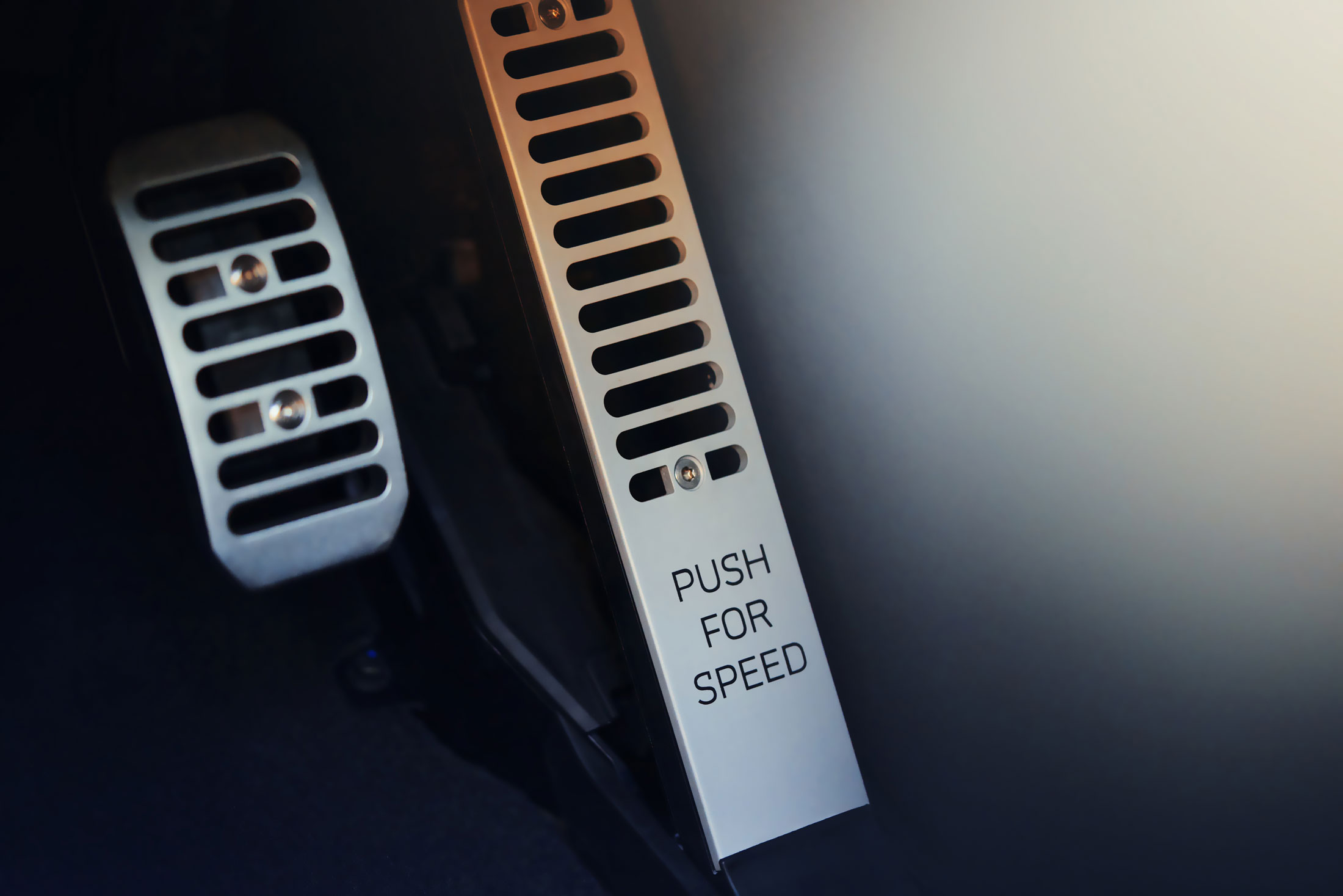 A close-up of a McLaren's metallic accelerator pedal, with the text 'PUSH FOR SPEED' engraved on it, alongside the brake pedal.