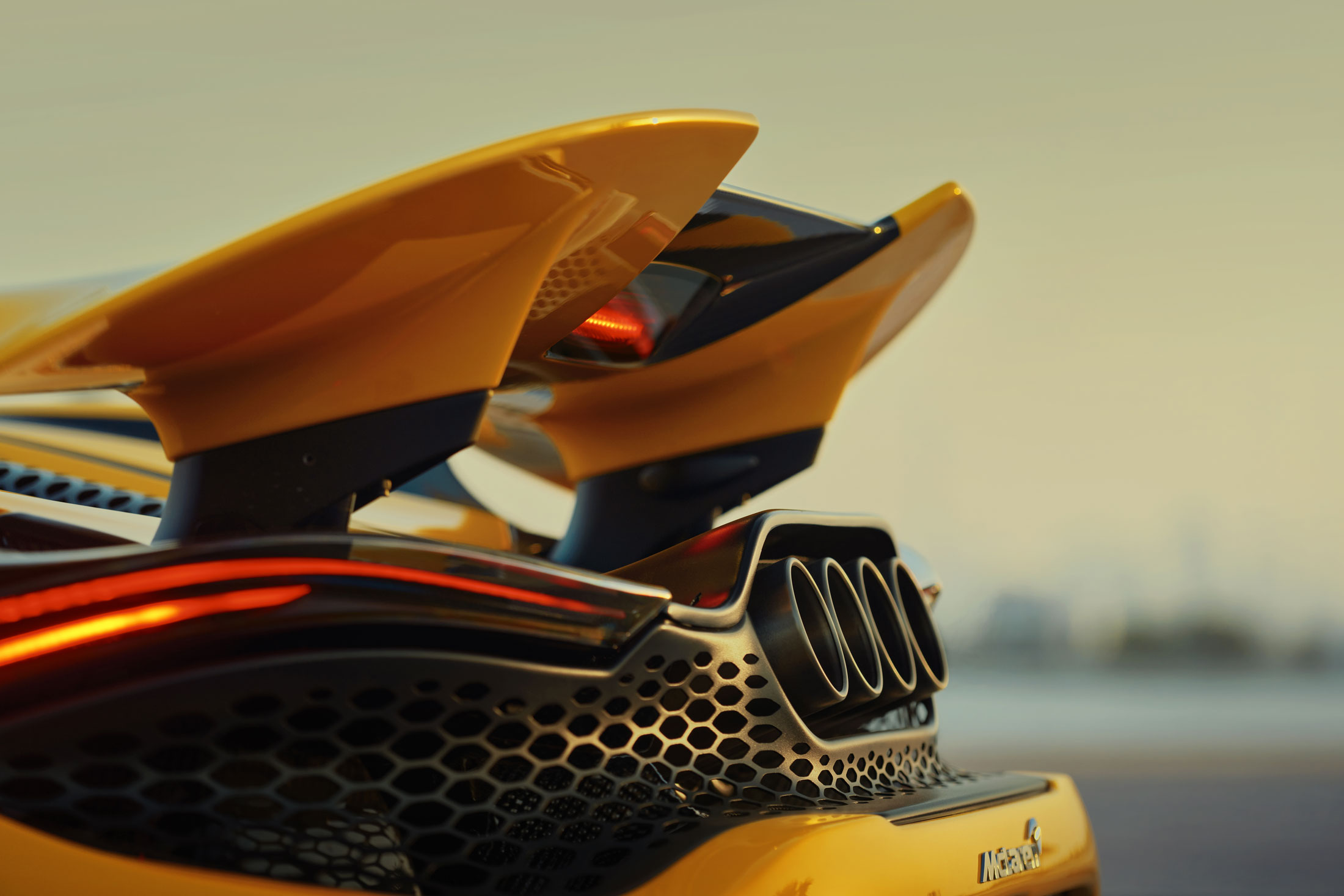 A close-up rear view of a yellow McLaren 765LT, highlighting the large active spoiler, the distinctive quad exhaust pipes, and the curved taillights.