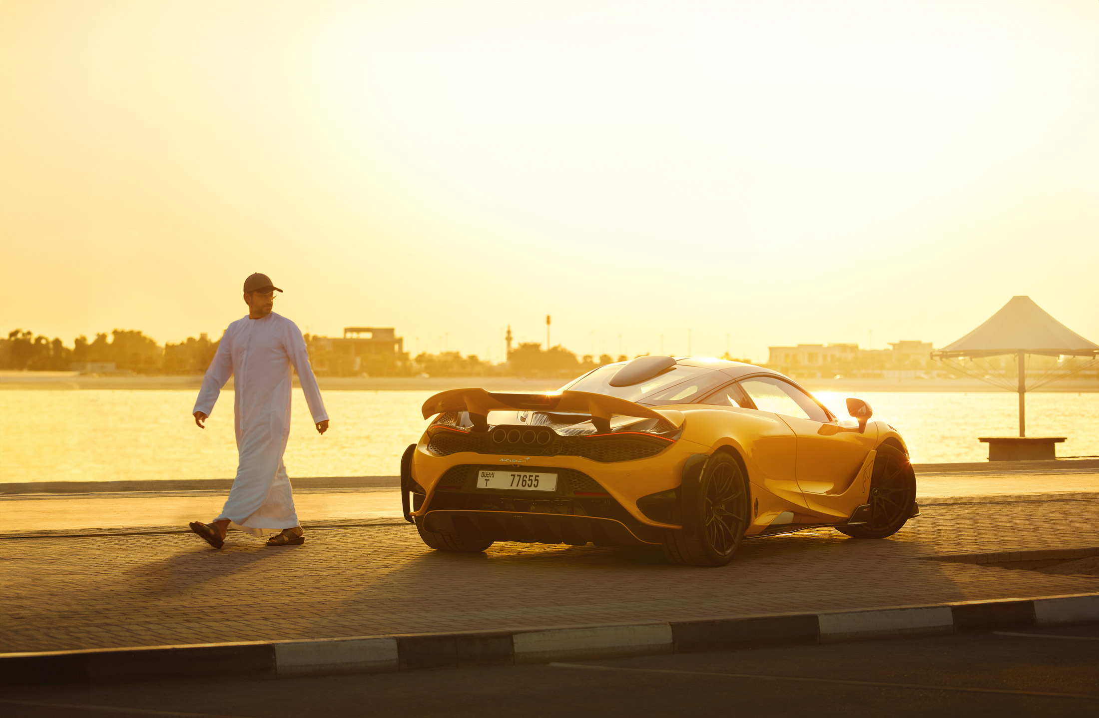 A striking rear-three-quarter view of a yellow McLaren 765LT parked by a calm body of water at sunset, with a man walking away from the car.