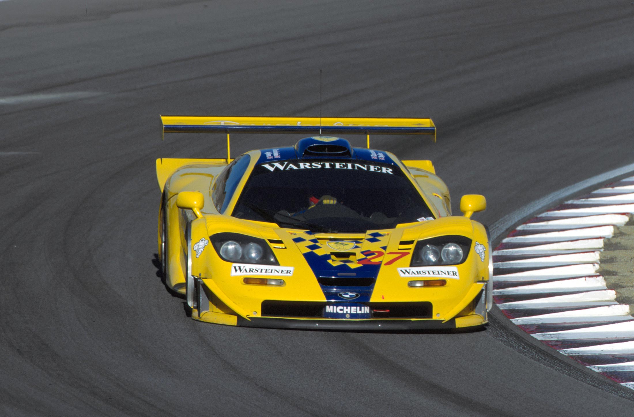 A professional action shot of a yellow McLaren F1 GTR 'Longtail' race car with blue accents, cornering on a racetrack.
