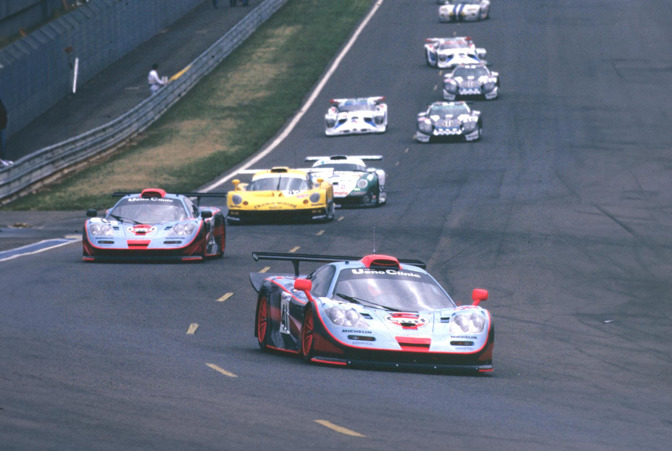 A vintage photo of a pack of McLaren F1 GTR 'Longtail' race cars racing down a track, with the race-winning FINA-liveried car in the foreground.