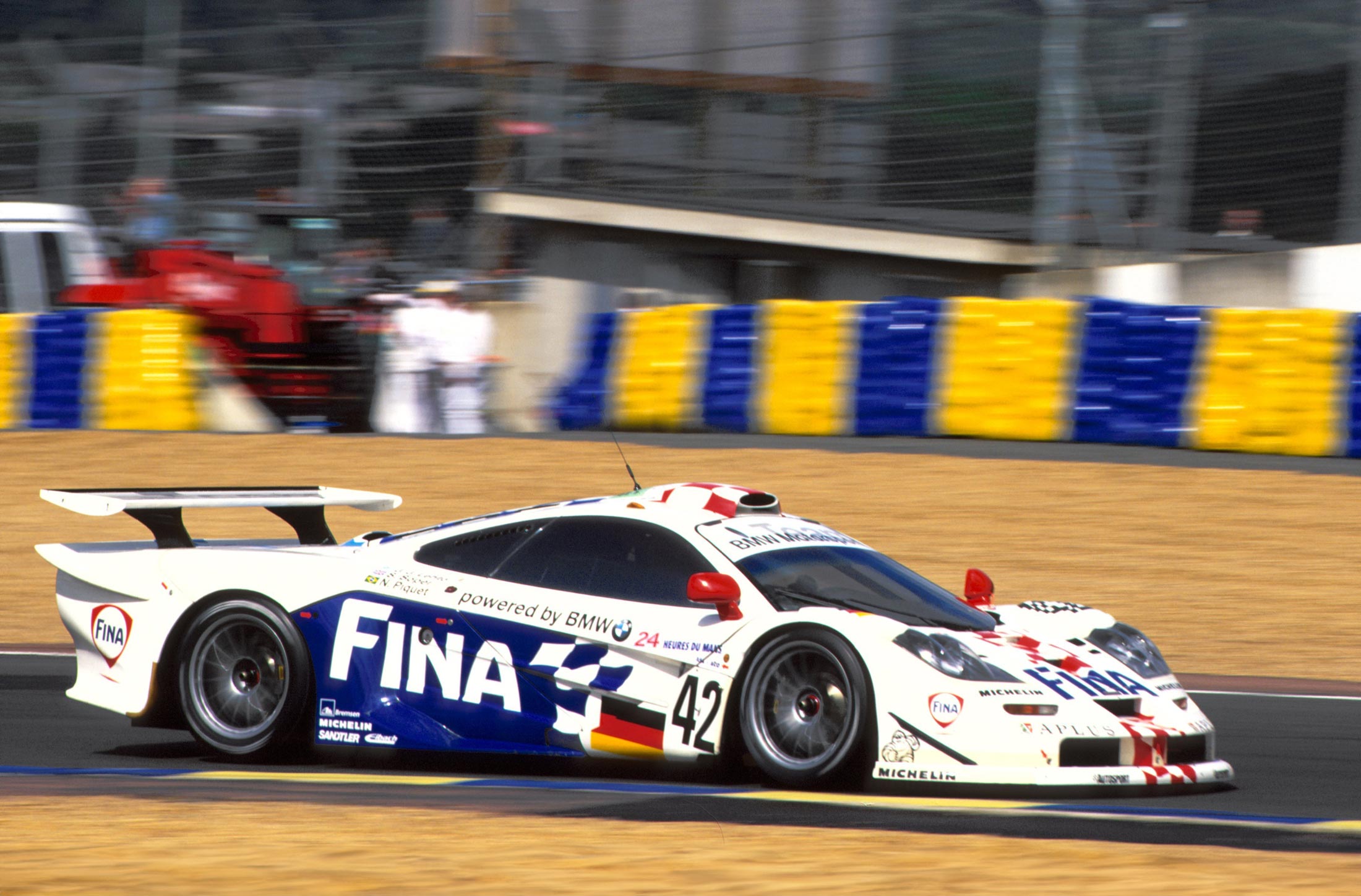 A vintage photo of a white, blue, and red McLaren F1 GTR 'Longtail' race car with the number 42 on a racetrack, with a red and blue striped wall in the background.