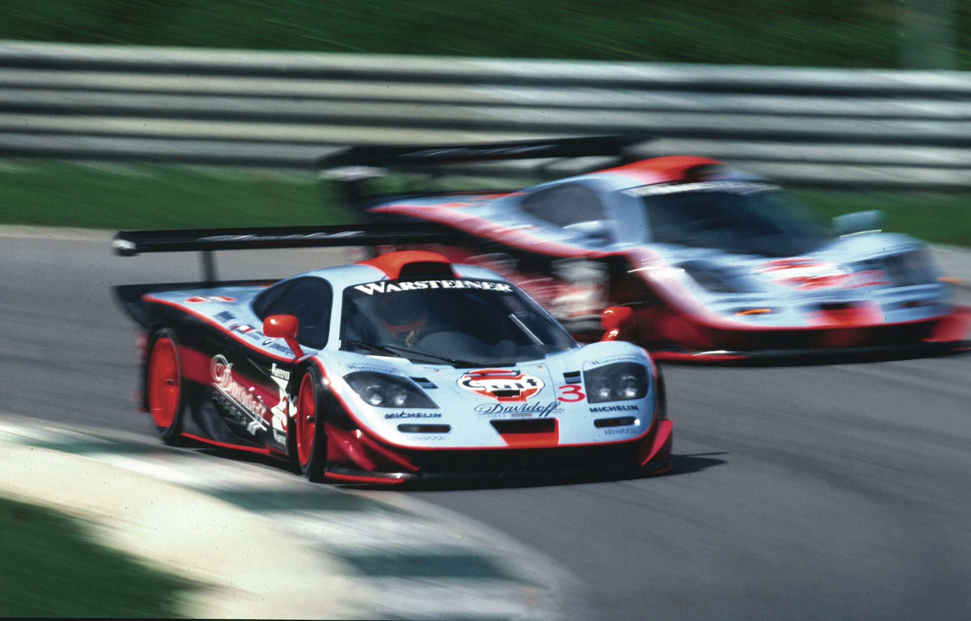 An action shot of two McLaren F1 GTR 'Longtail' race cars with a red, white, and blue livery racing on a track with motion blur.