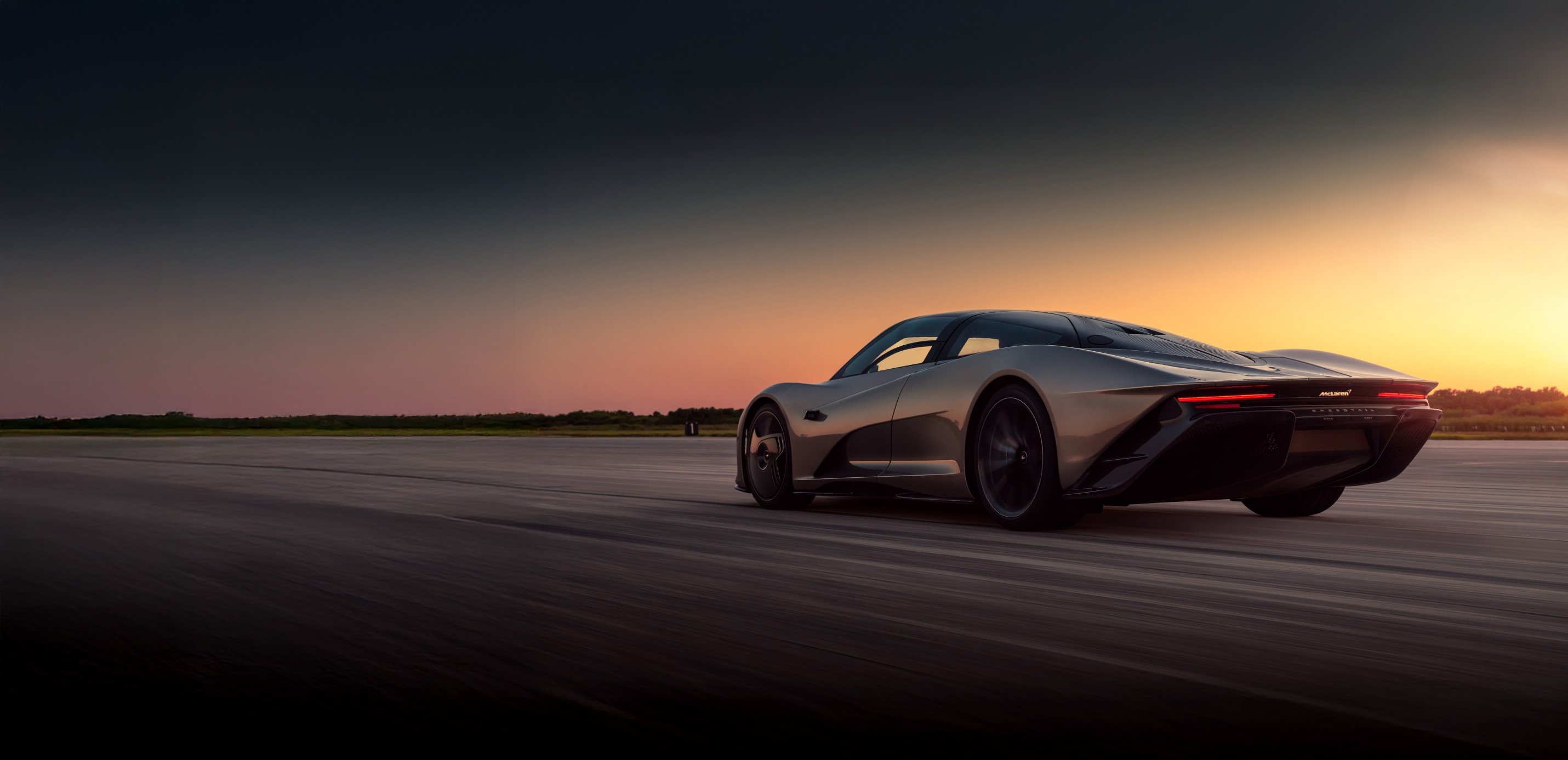 Grey McLaren Speedtail driving down a road at speed during sunset. The colours of the sky reflecting off of the car body.