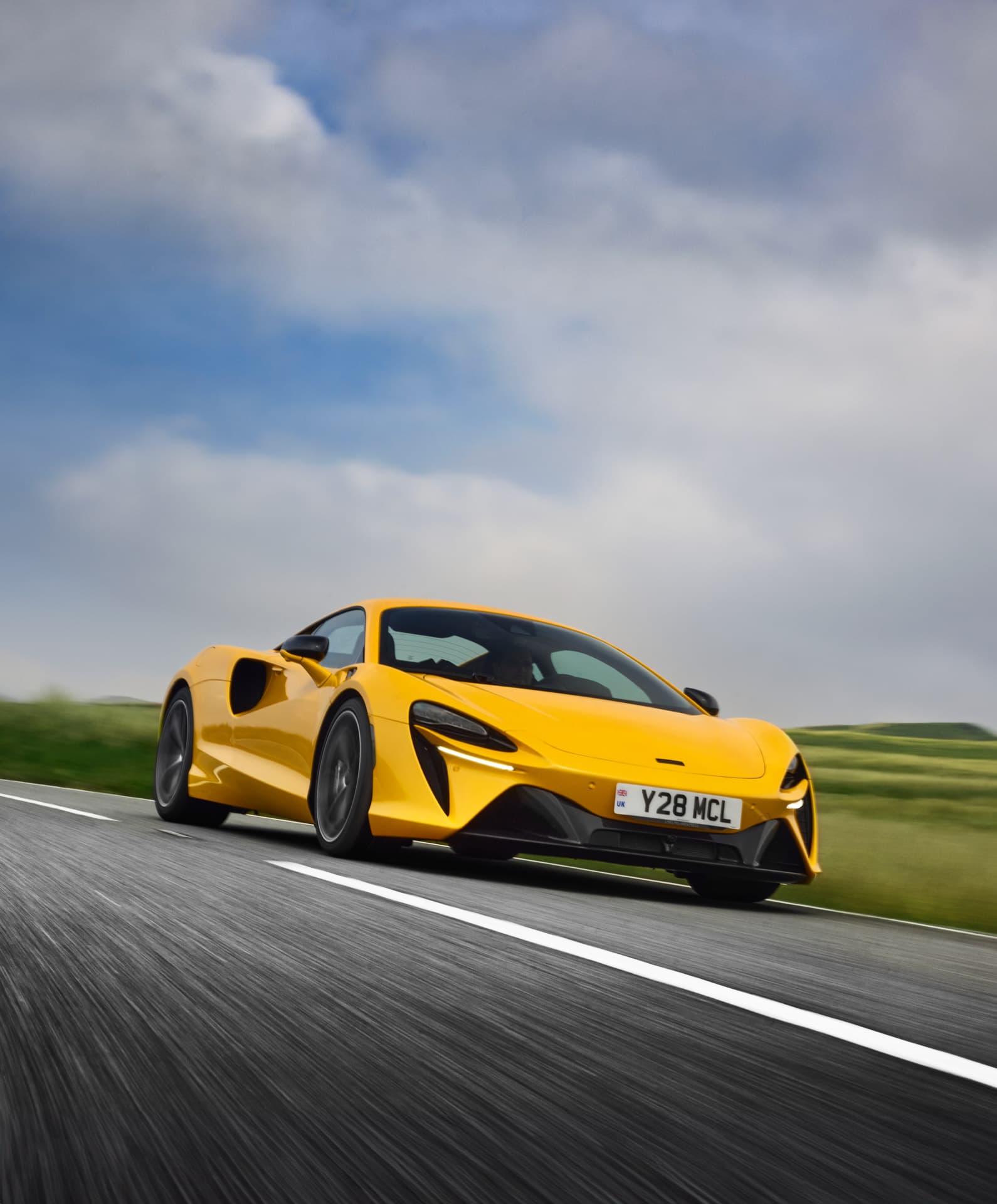 A professional action shot of a yellow McLaren Artura from a front-three-quarter view, speeding on a country road with a blurred background.