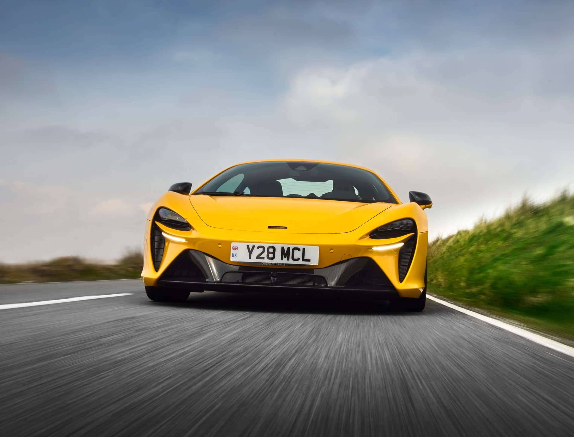 A dynamic low-angle shot of a vibrant yellow McLaren Artura supercar driving on a road, emphasizing its sleek front design against a motion-blurred landscape.