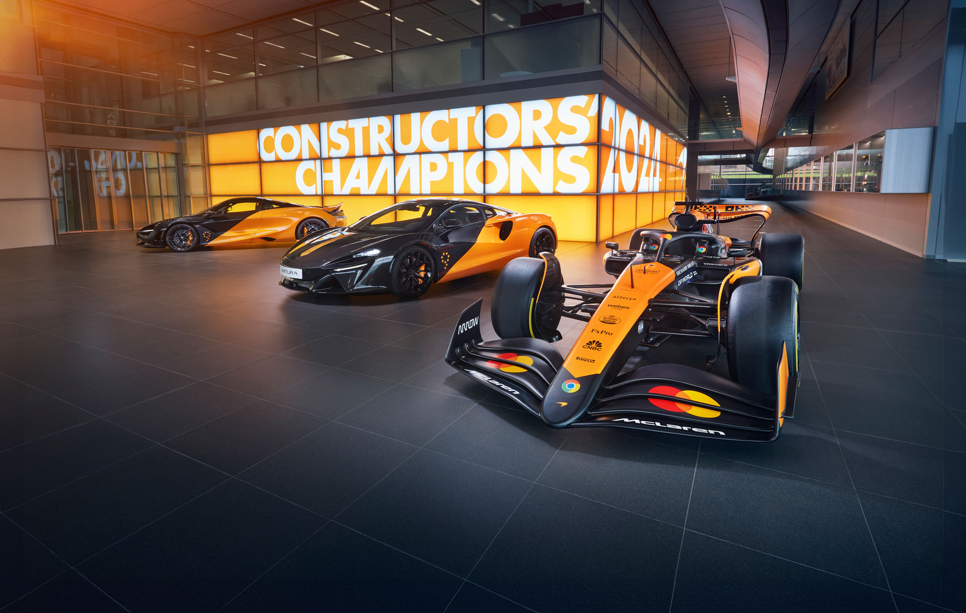 A hero shot of a McLaren Formula 1 car, a McLaren Artura, and another McLaren supercar, all in the iconic orange and black livery, displayed in a showroom.