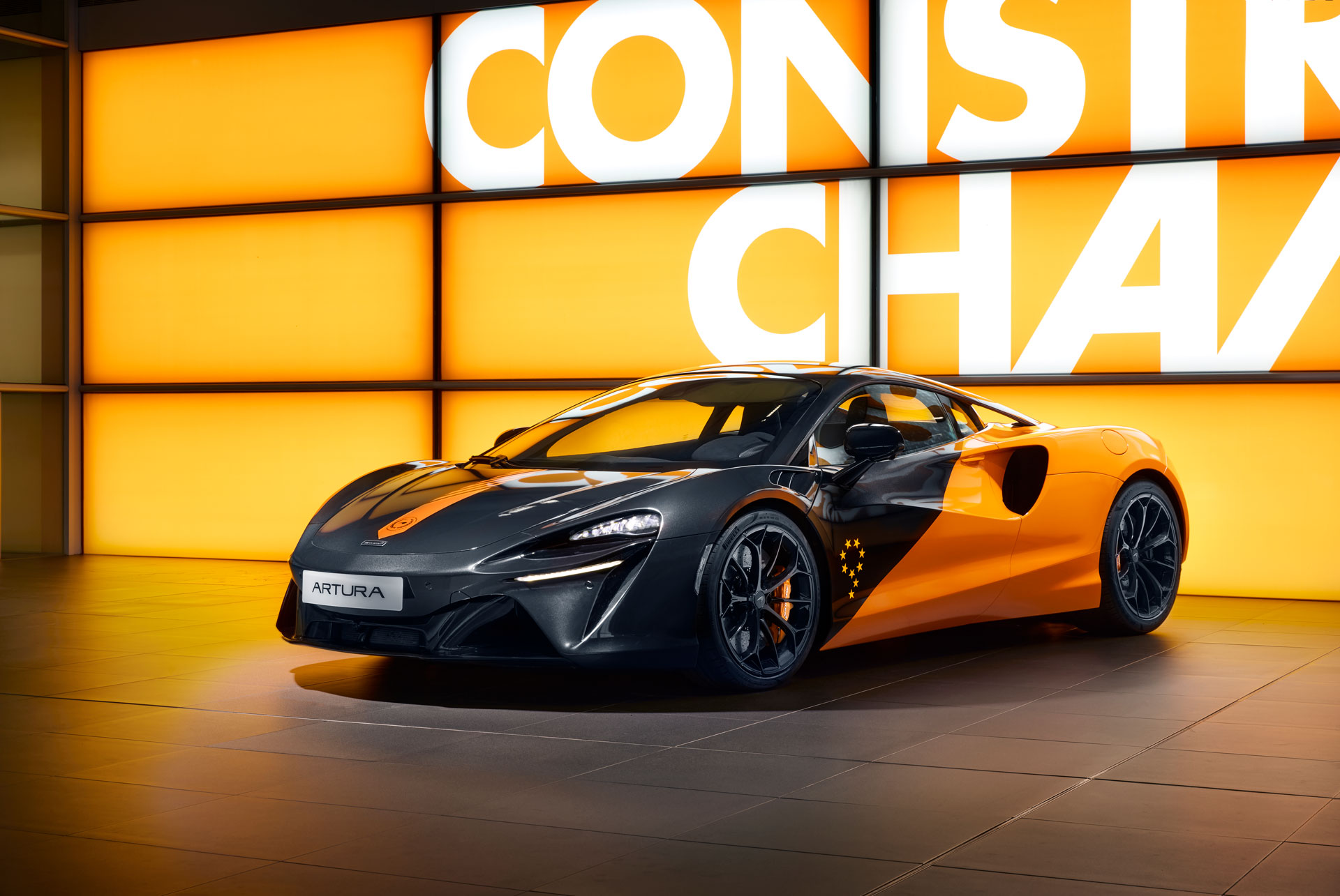 A front-three-quarter view of a black and orange McLaren Artura supercar, showcasing its custom livery in a showroom with a 'CONSTRUCTORS' CHAMPIONS' wall in the background