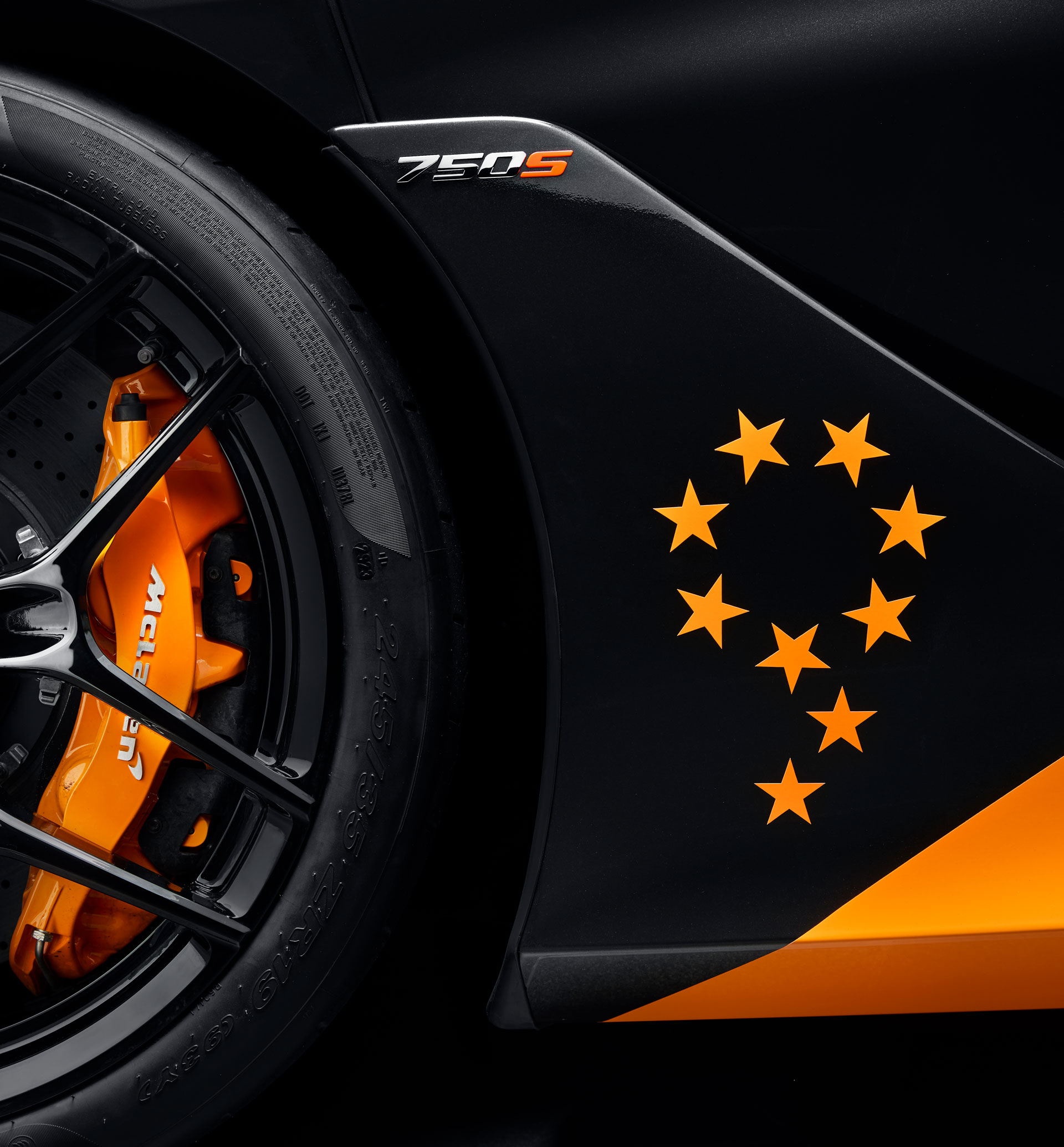 A close-up of a black McLaren 750S's side, highlighting the orange brake caliper and a unique graphic of nine stars arranged in the shape of the number nine.