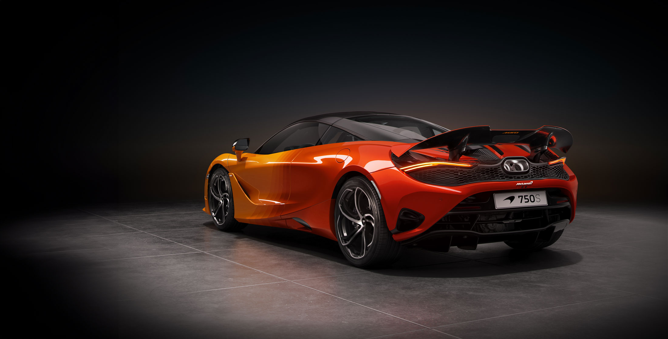 A stunning rear-three-quarter view of a McLaren 750S supercar with a custom gradient livery that fades from yellow to orange, highlighting its active rear spoiler and glowing taillights in a studio setting.