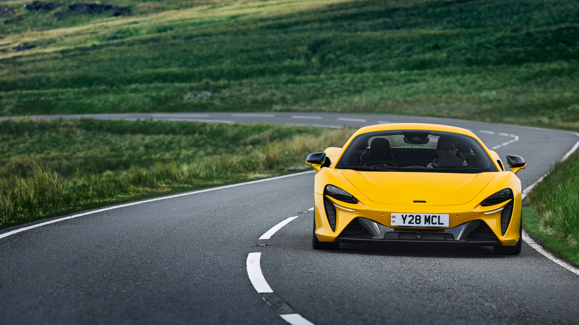 A vibrant yellow McLaren Artura supercar drives on a winding road through a green, hilly landscape, captured in a front-on view.