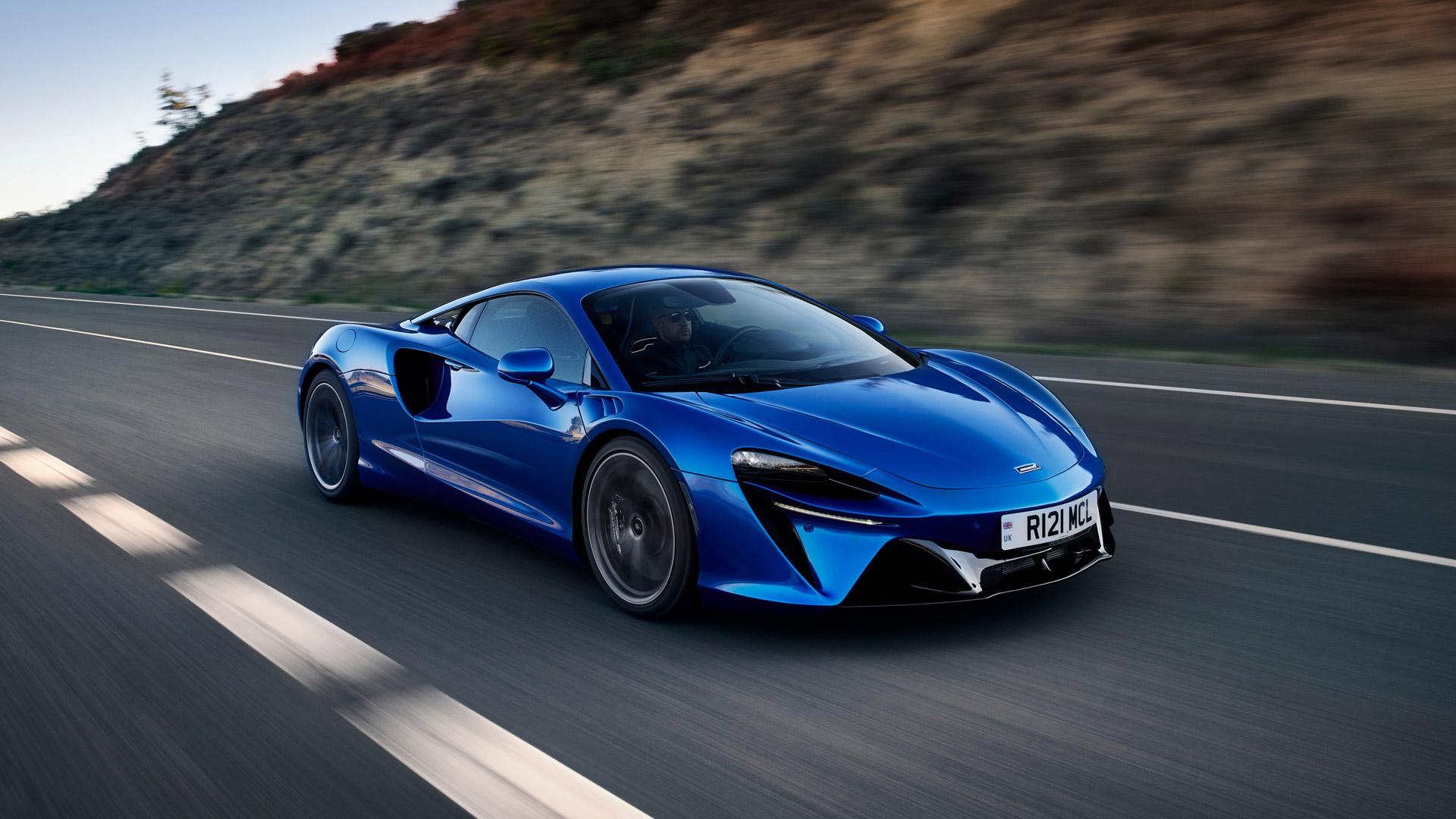The McLaren Artura hybrid supercar in a brilliant blue colour, captured in a high-speed motion shot on a highway.