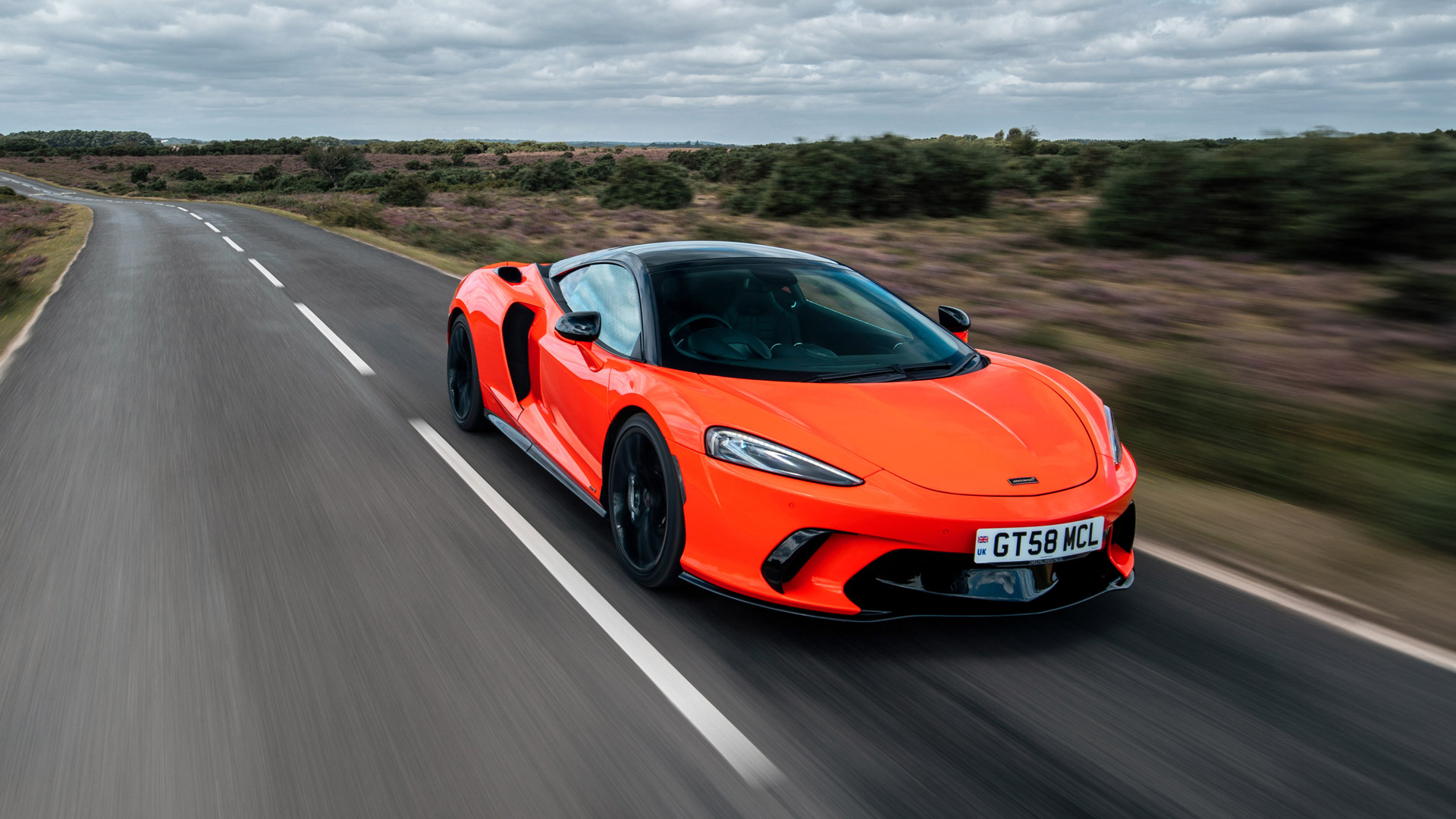 The McLaren GTS in a bright red finish driving on a twisting road, surrounded by countryside.