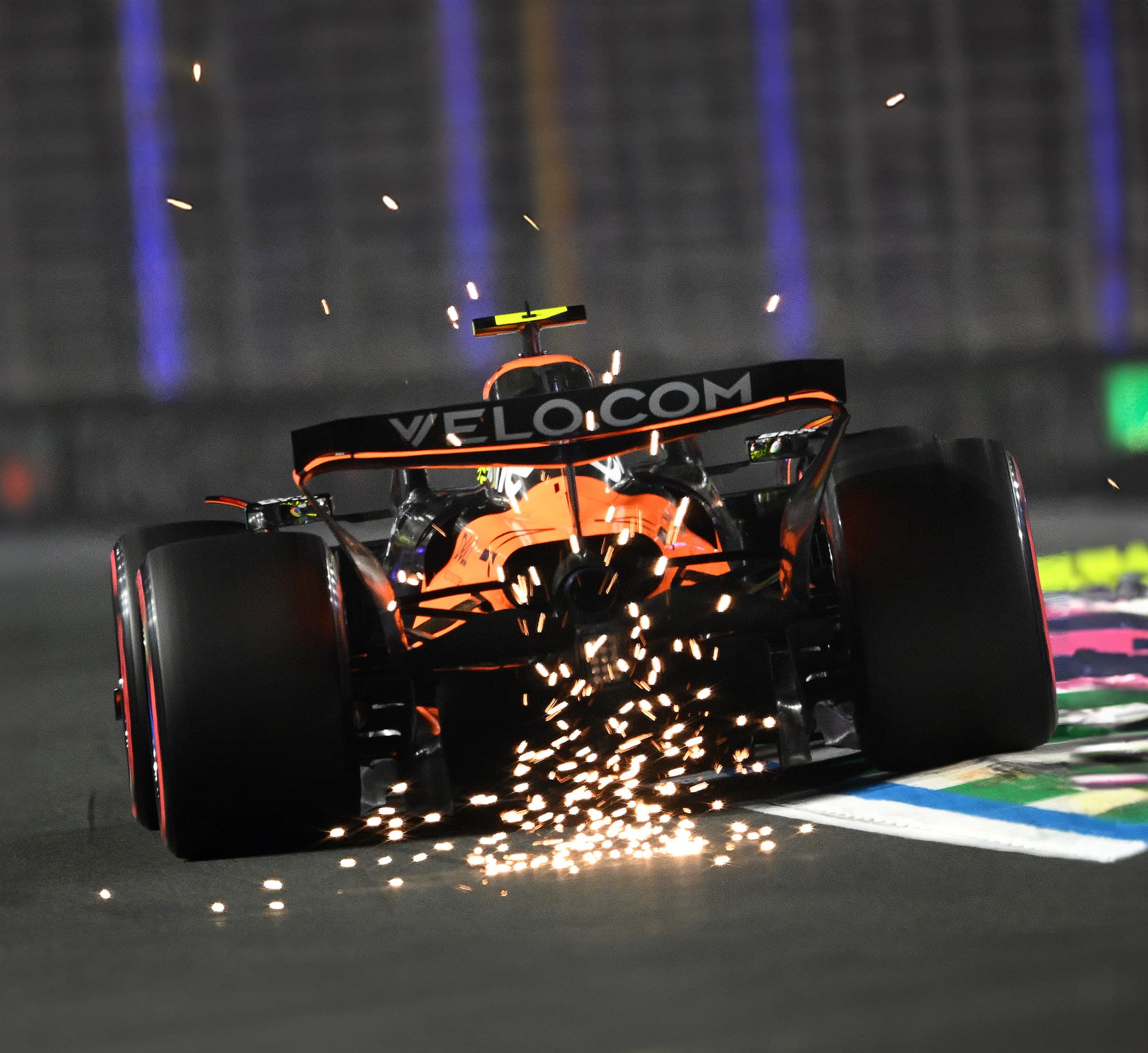 The rear of the McLaren MCL39 F1 car kicking up a shower of sparks at high speed on the track at night.