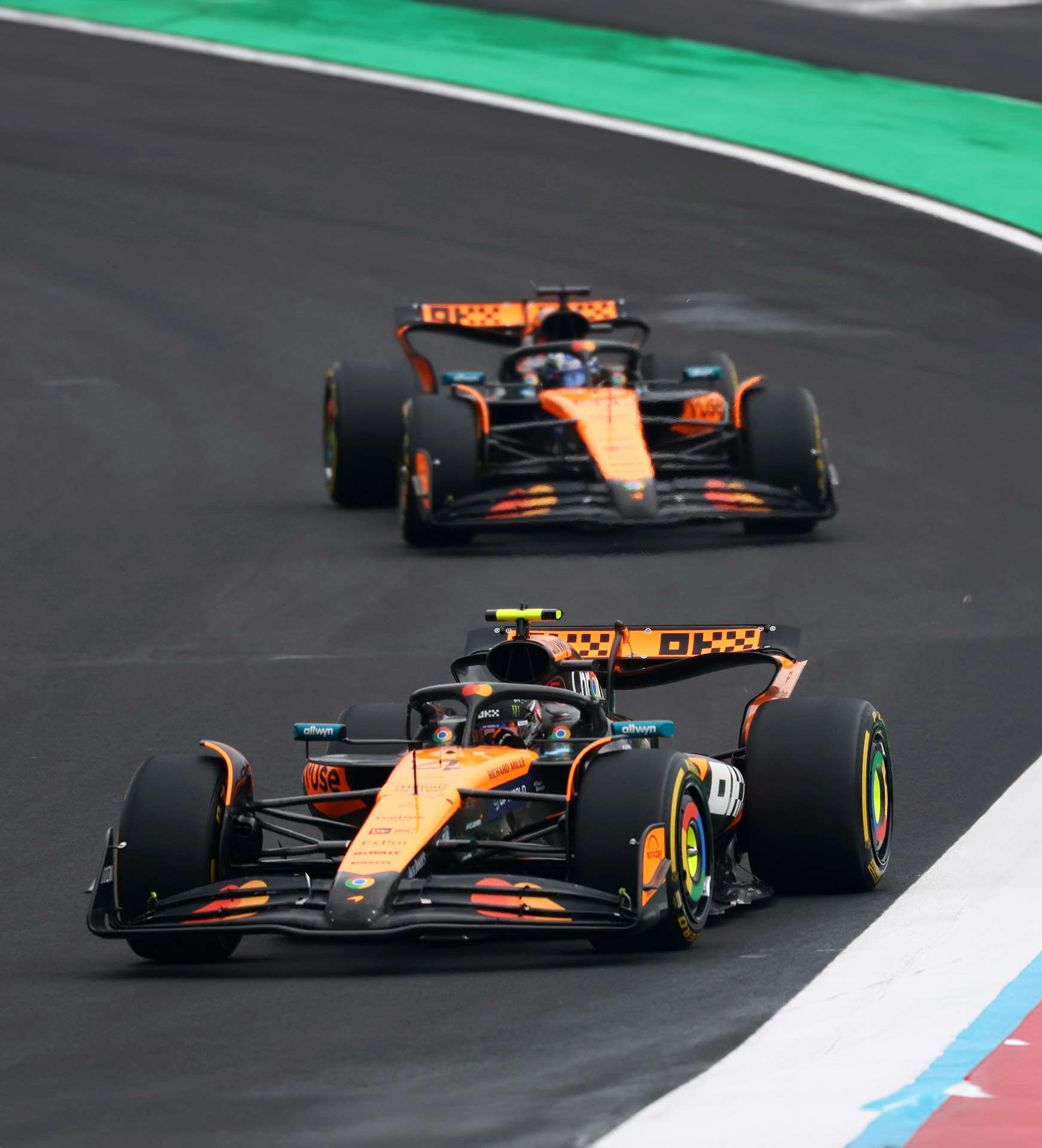 Two McLaren F1 cars, the MCL39, racing in formation on the circuit, highlighting the team's iconic papaya and black livery.
