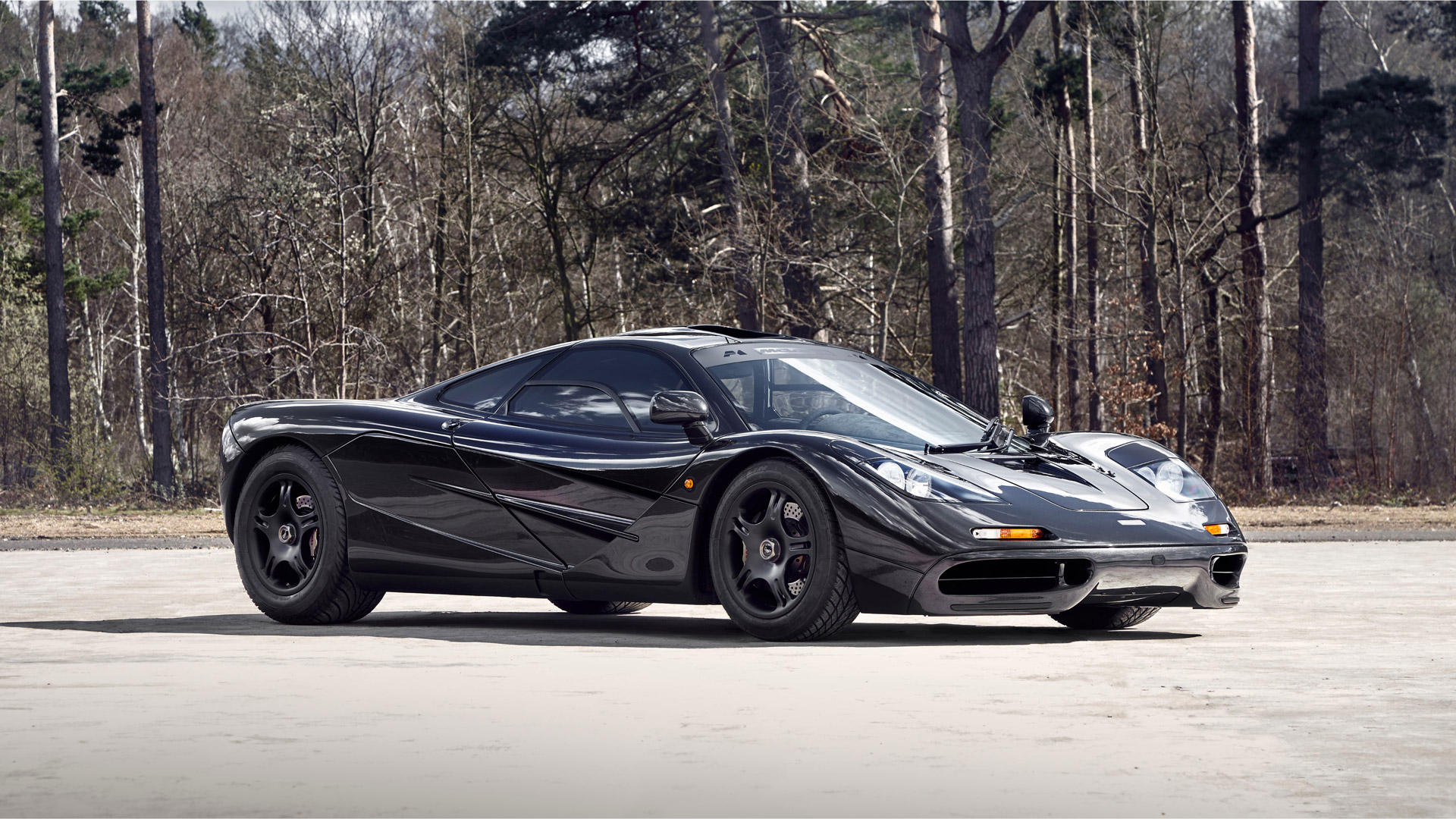A sleek black McLaren F1 supercar with its distinct central driving position visible, parked in a wooded area on a sunny day.