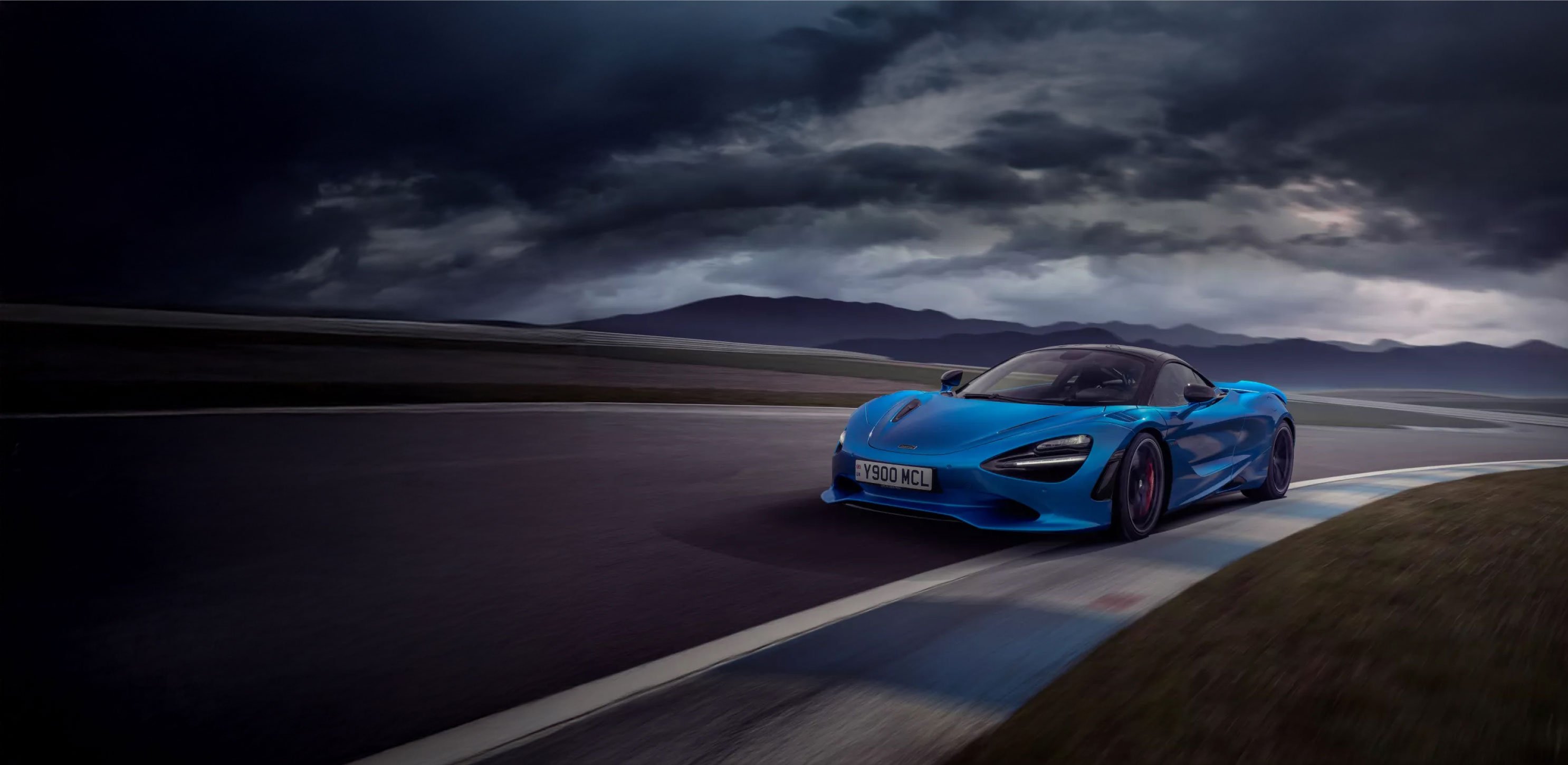 An action shot of a bright metallic blue McLaren 750S driving on a racetrack. The car is turning a corner, with its wheels visible and a sense of motion blur on the road surface.
