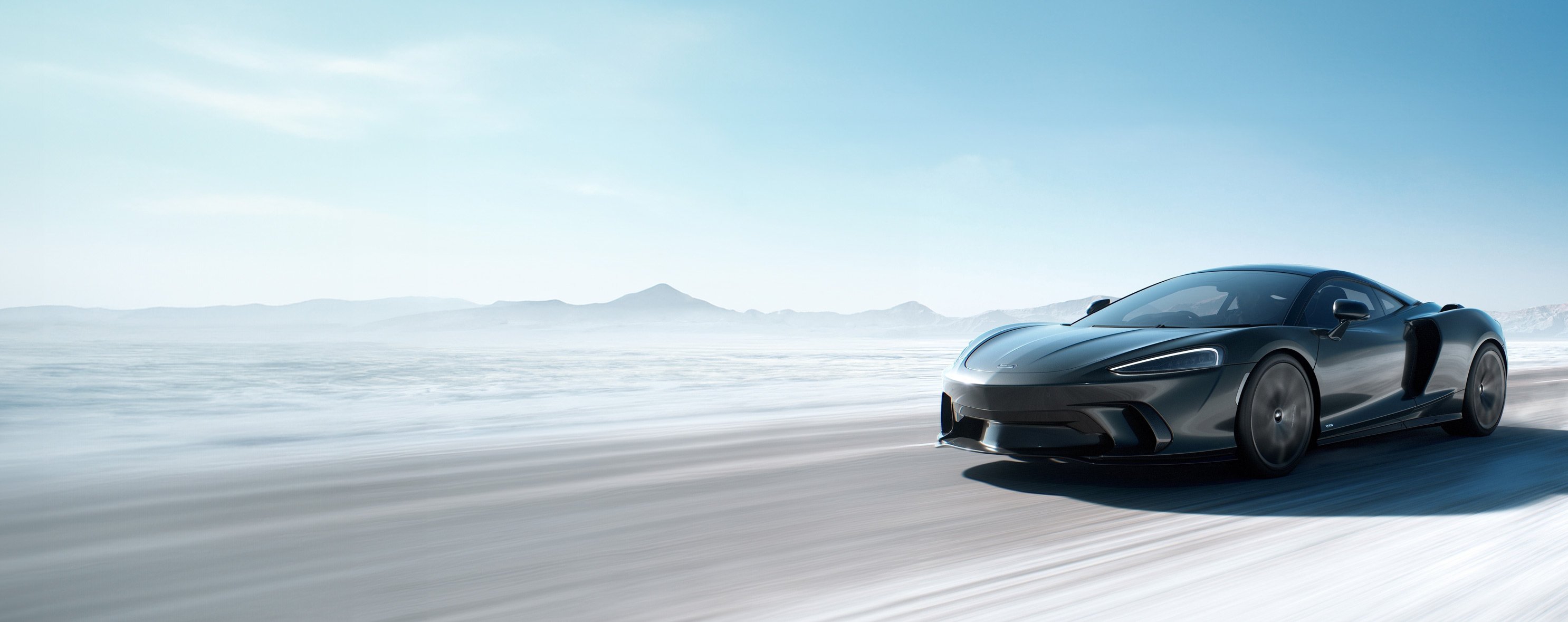  A dark grey McLaren GT drives on a vast, flat, snowy landscape with distant mountains under a bright, clear sky, conveying speed and open space.