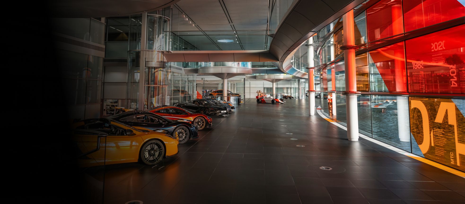 Inside the McLaren headquarters. On the left there is a row of McLaren cars lined up on display. Then to the left there are floor to ceiling windows that overlook the McLaren lake.