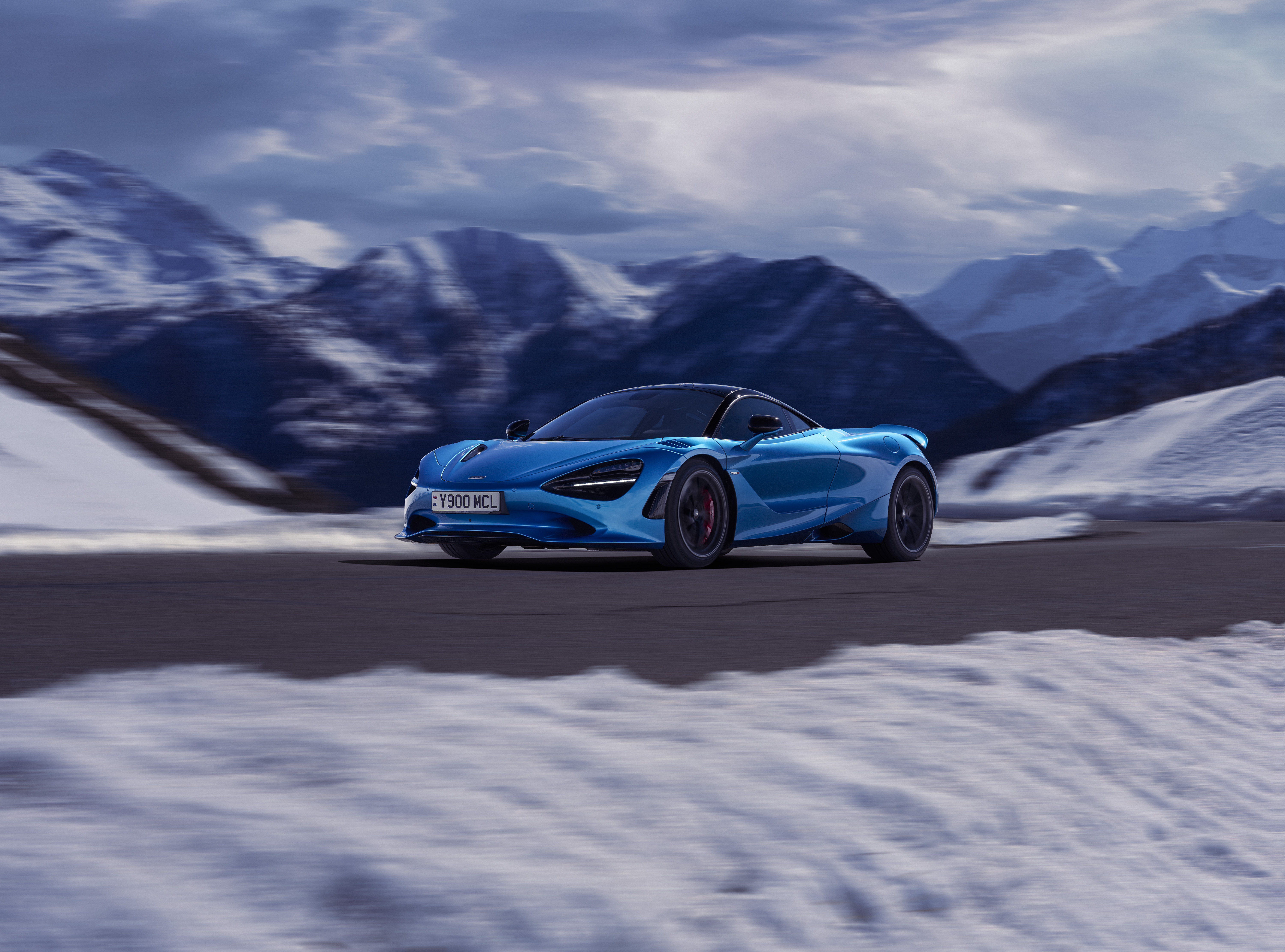 Blue McLaren 750S driving down a snowy mountain road at speed, on a cloudy day.