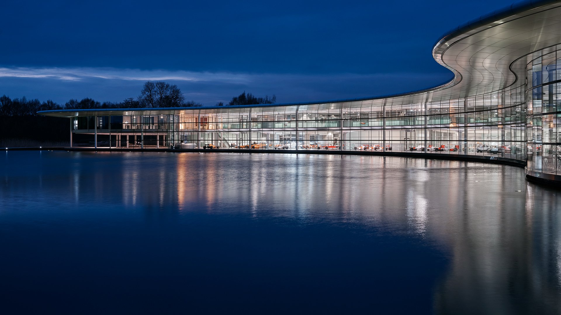 The iconic McLaren Technology Centre in Woking, England, illuminated at dusk with the building's lights reflecting on the lake.