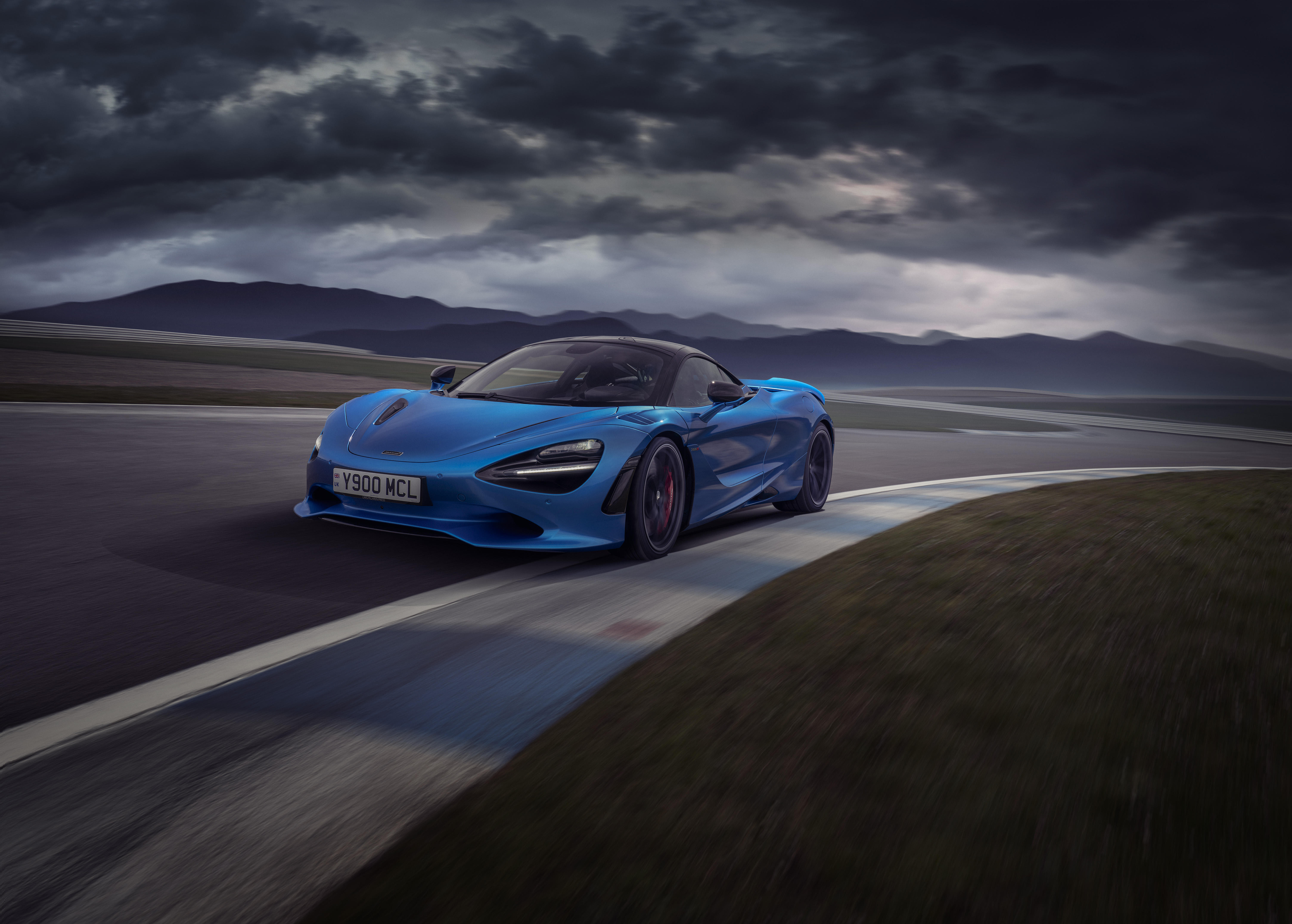 Front view of blue McLaren 750S driving around a race track at speed, on a cloudy day.