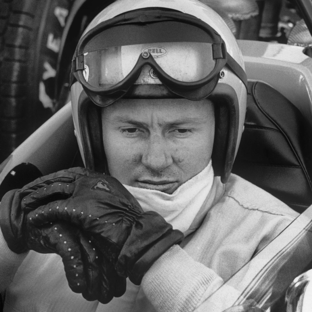 Bruce McLaren sat in a race car wearing a helmet and driving gloves.