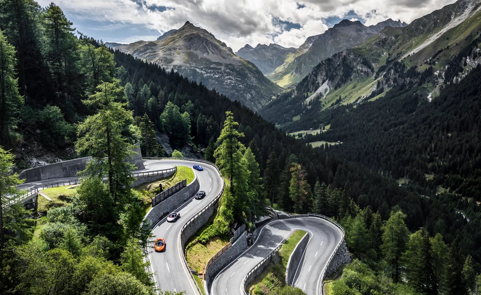A convoy of McLaren sports cars in various colors navigating a dramatic, winding mountain pass, showcasing the ultimate grand touring experience.