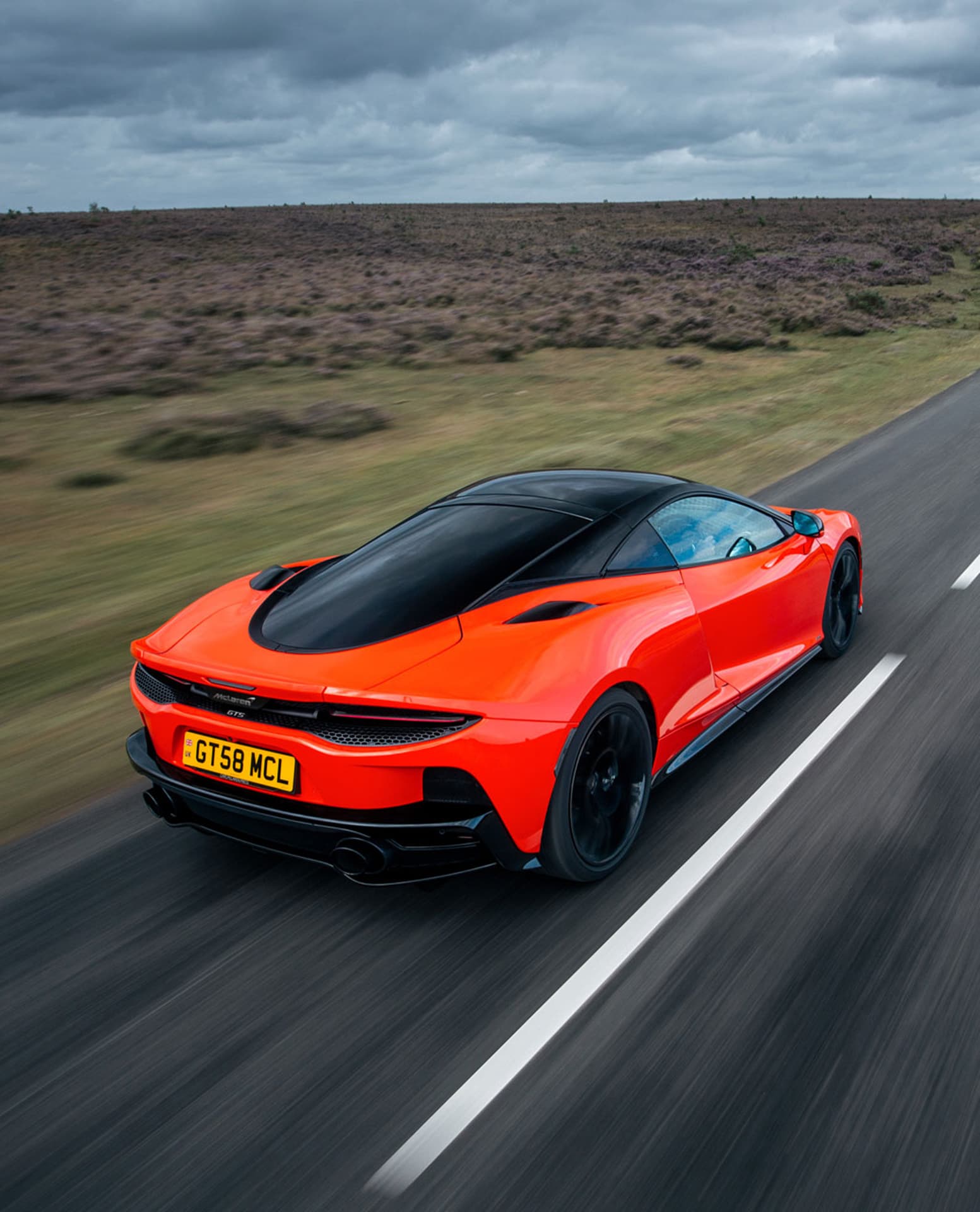 A high-angle rear view of a bright red McLaren GTS speeding down a winding road, with motion blur indicating speed, set against a wide, somewhat barren landscape under a cloudy sky.