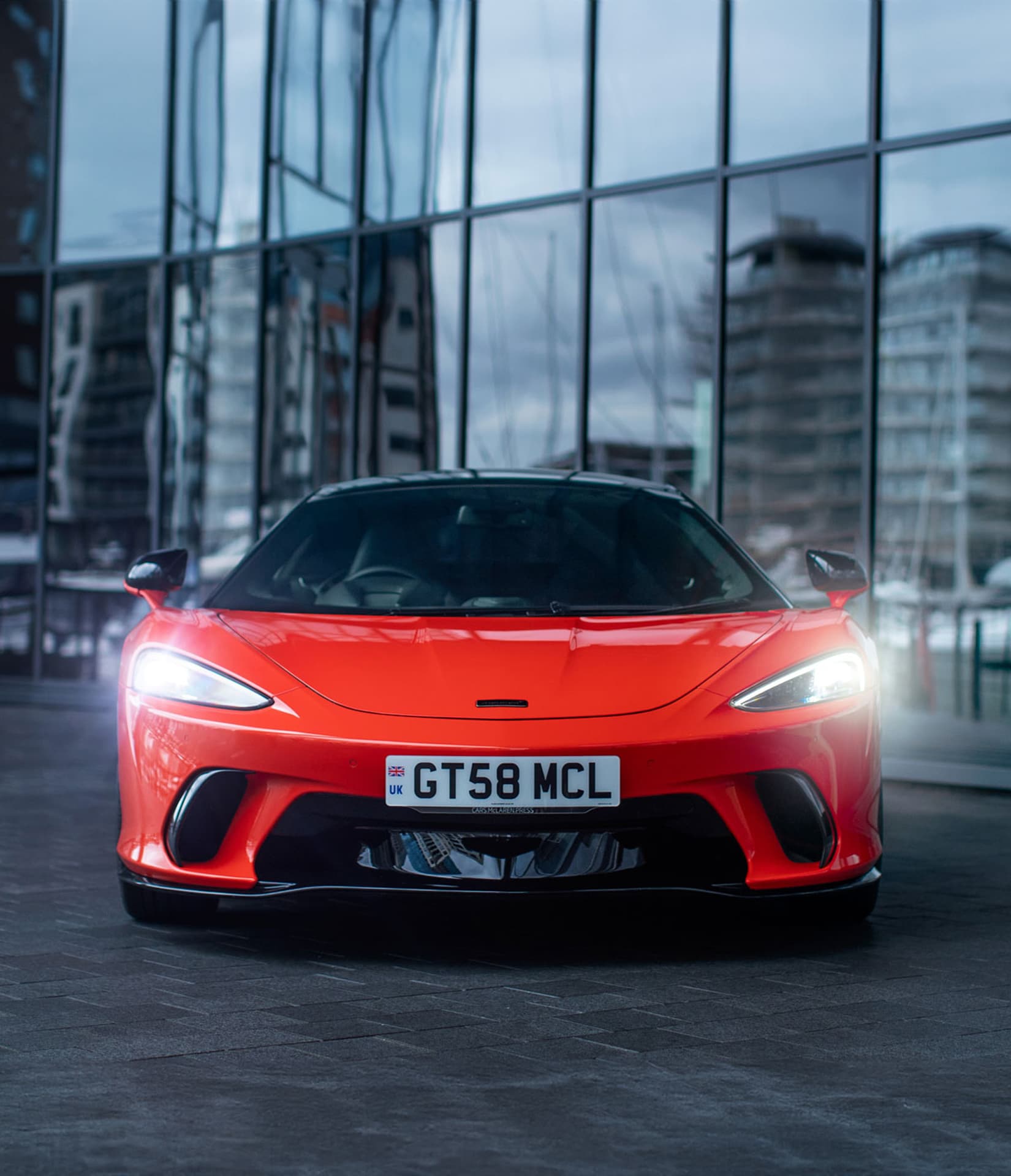 A bright red McLaren GTS with its headlights on is parked directly facing the viewer in front of a modern building with large reflective glass windows.