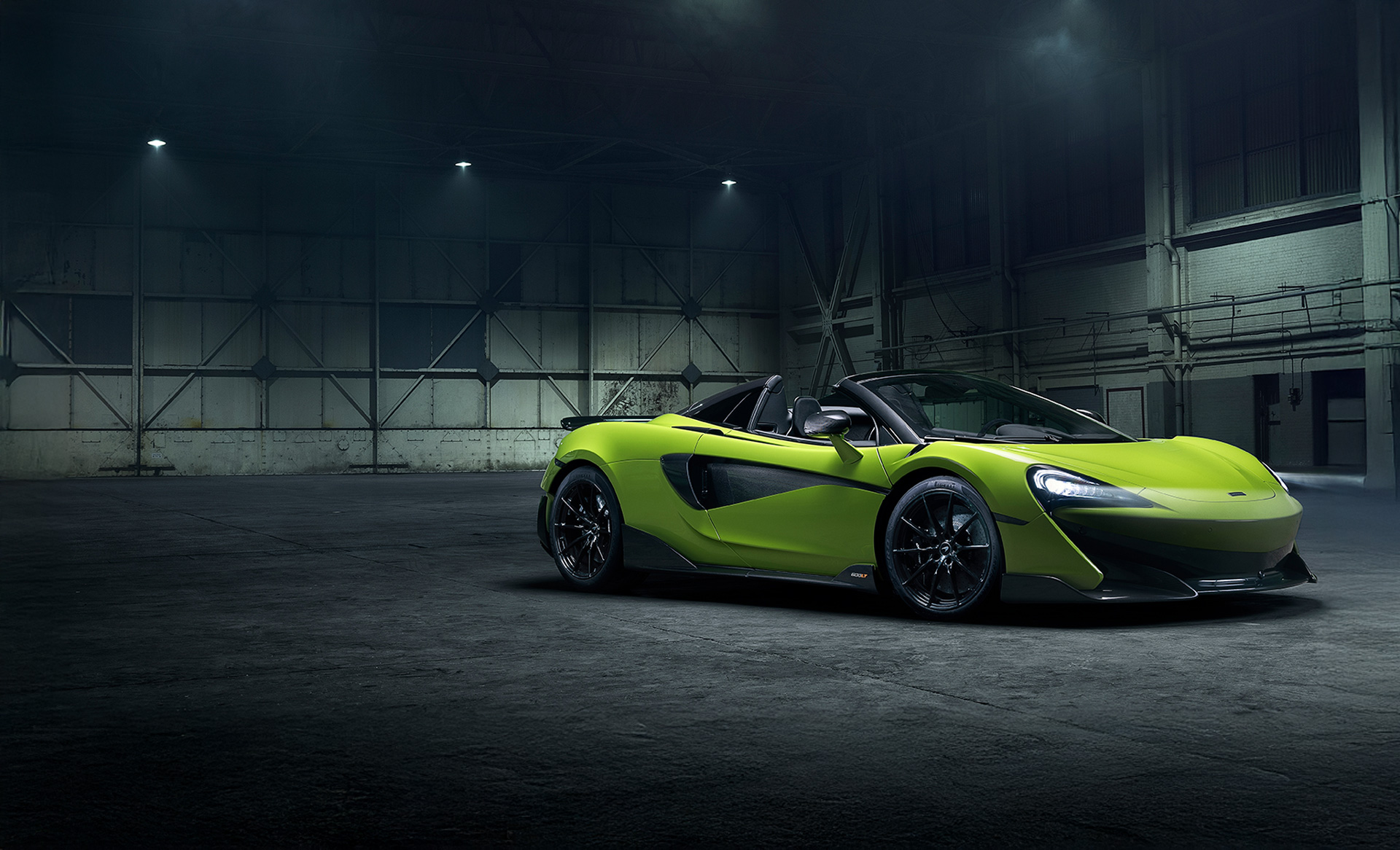 A studio shot of a McLaren 600LT Spider convertible. The car is painted in a vibrant, bright lime green and is parked indoors in a large, dark, industrial setting