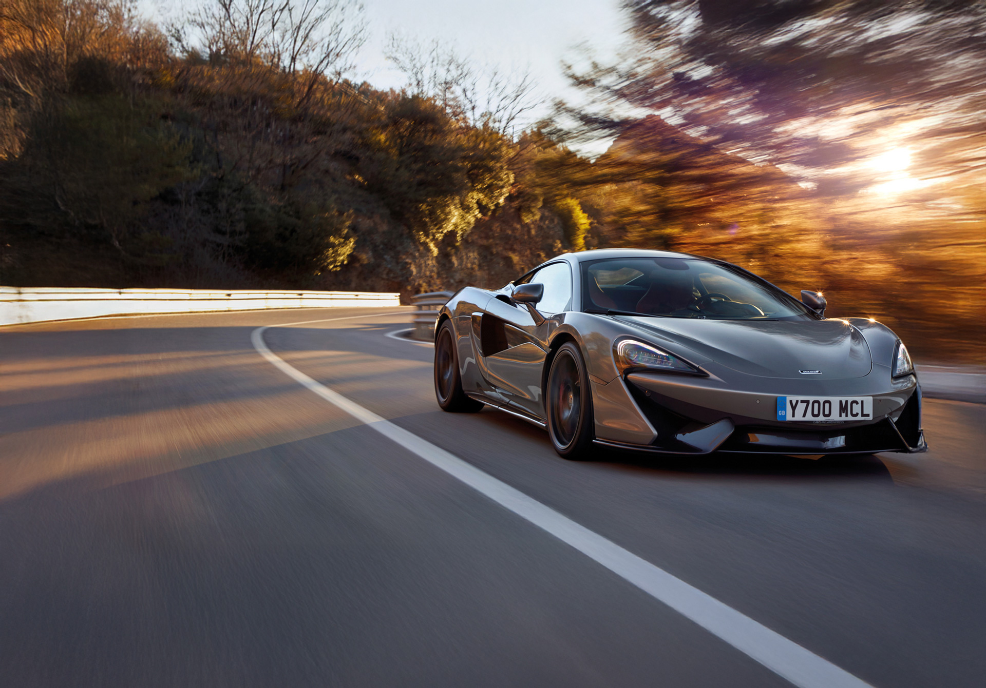 A grey McLaren 570S driving down a road at speed, surrounded by autumnal tree, in golden sunlight. The reflection of the trees is visible on the car body.