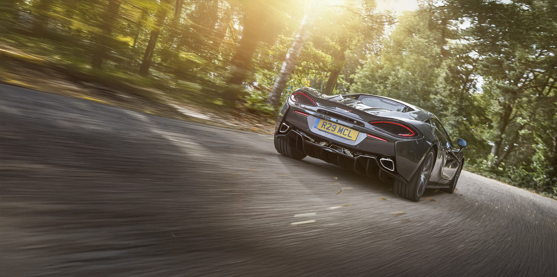 A rear low-angle shot of a dark grey McLaren supercar accelerating on a winding road, showcasing its distinctive taillights, exhaust pipes, and license plate, with sun-dappled trees in the background.