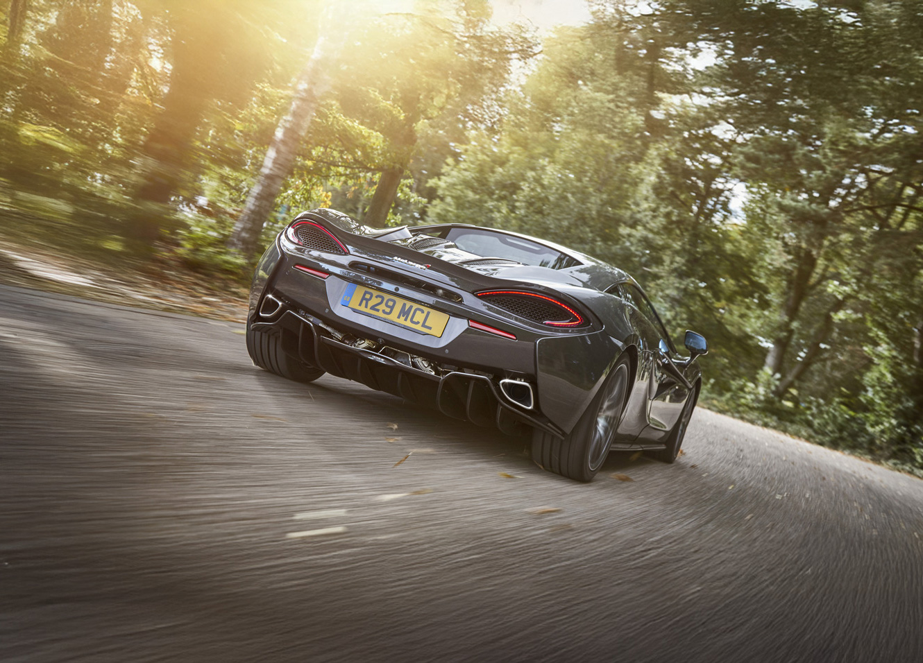 A dark grey McLaren 570GT supercar driving on a winding road with trees, seen from the rear.