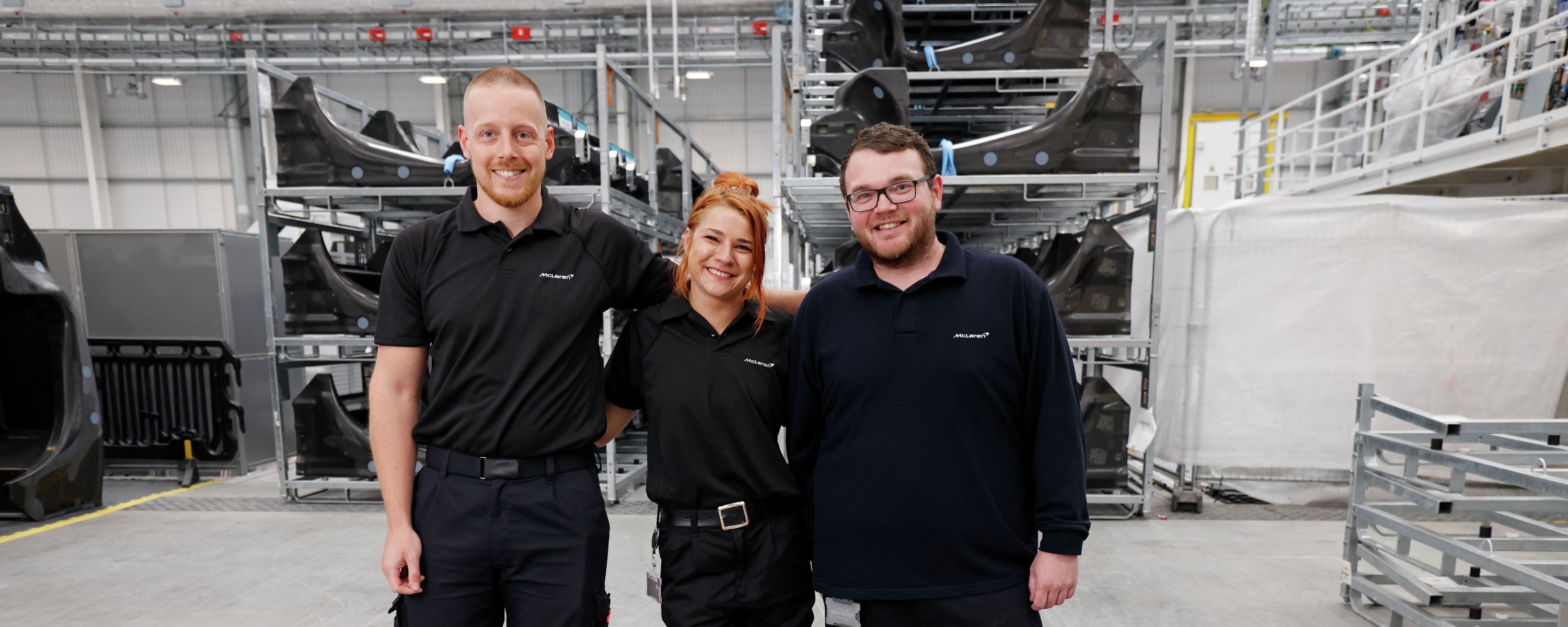  Three McLaren factory workers in uniform, standing and smiling inside a manufacturing facility.