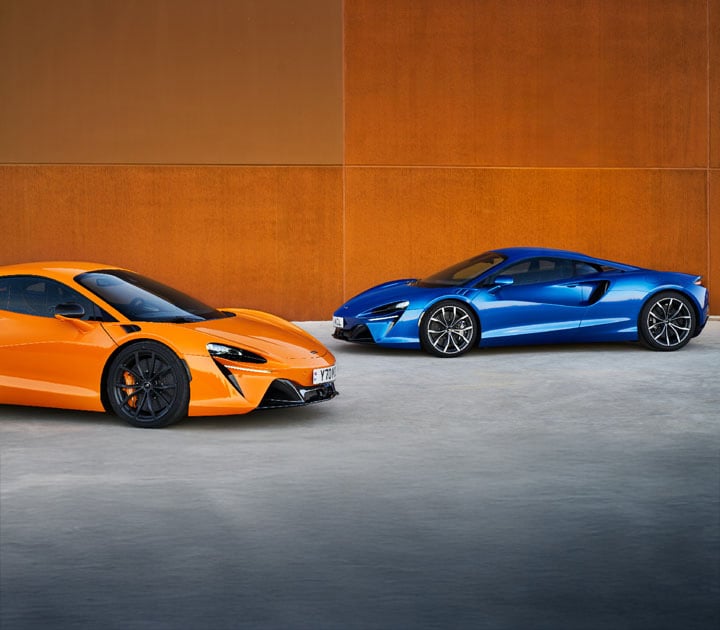 An orange and blue McLaren Artura parked in front of a rustic orange building.