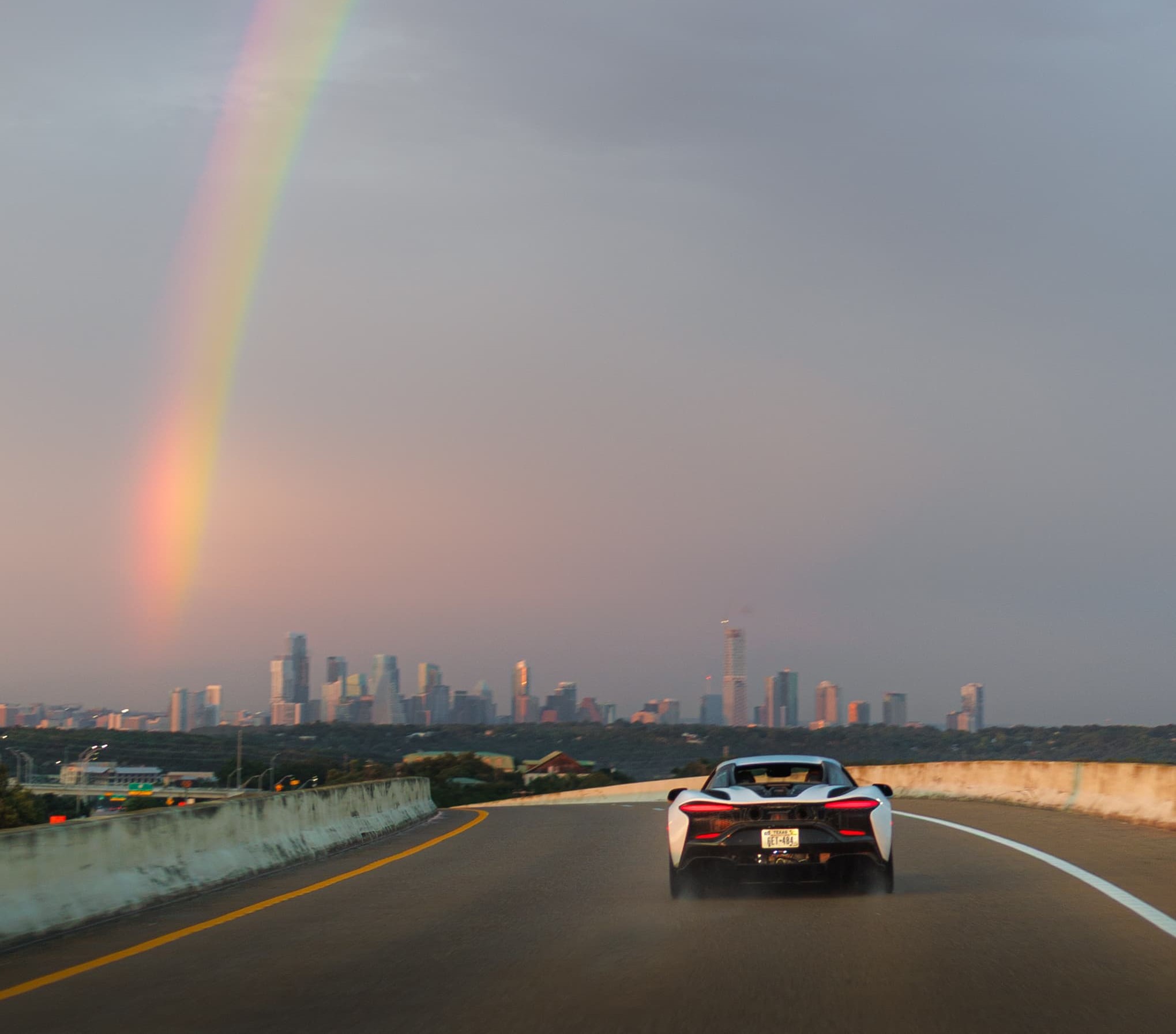 A white McLaren driving over a bridge towards Austin, Texas, at sunset, with a rainbow in the sky. A scene from Day 5 of the 'McLaren States of Endurance' road trip.