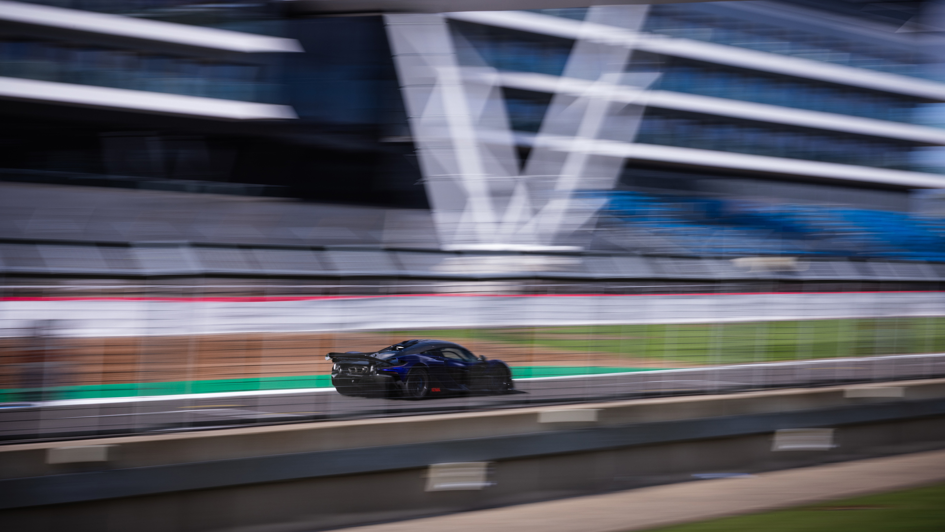 A shot of the rear side of a McLaren W1 prototype in royal blue driving at speed around the Silverstone track with motion blur to enhance the idea of speed.