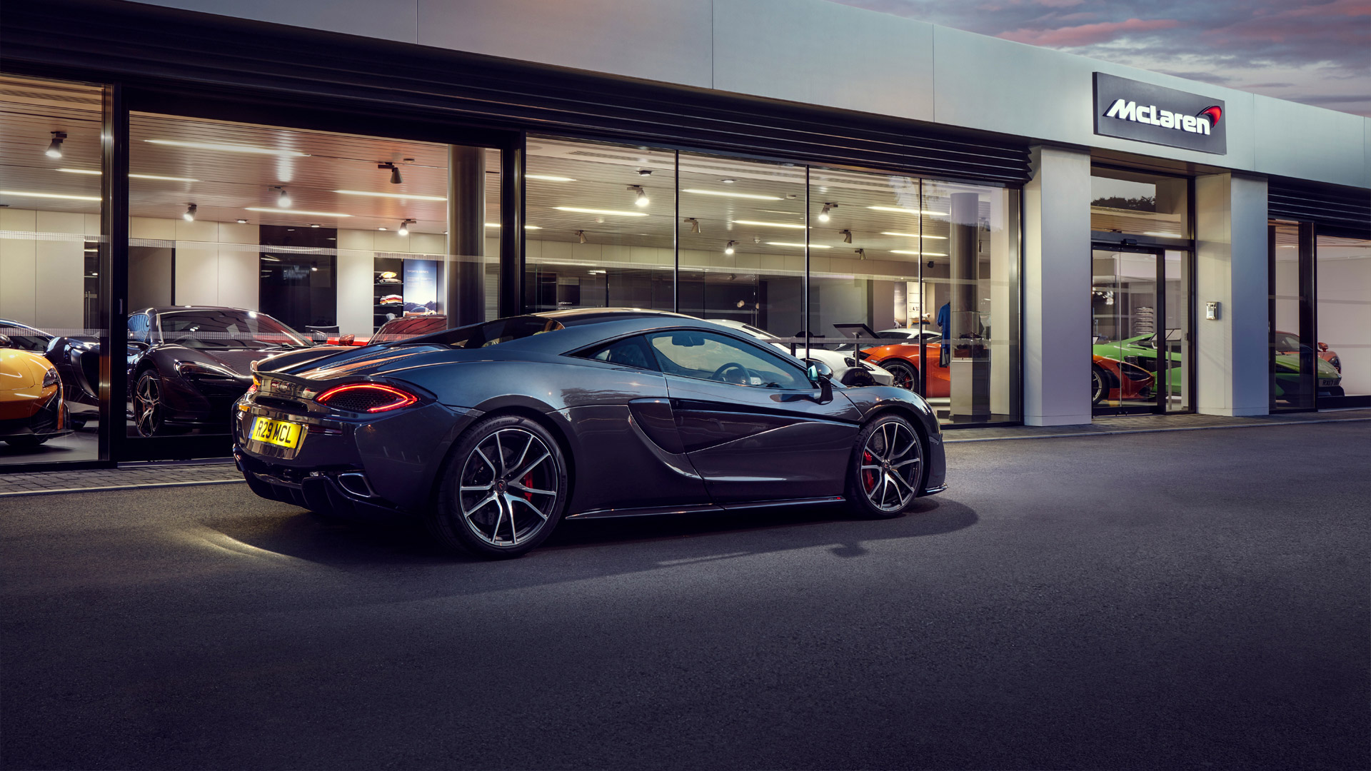 A sleek, dark grey McLaren sports car is parked on the asphalt in front of a modern McLaren retailer or dealership.